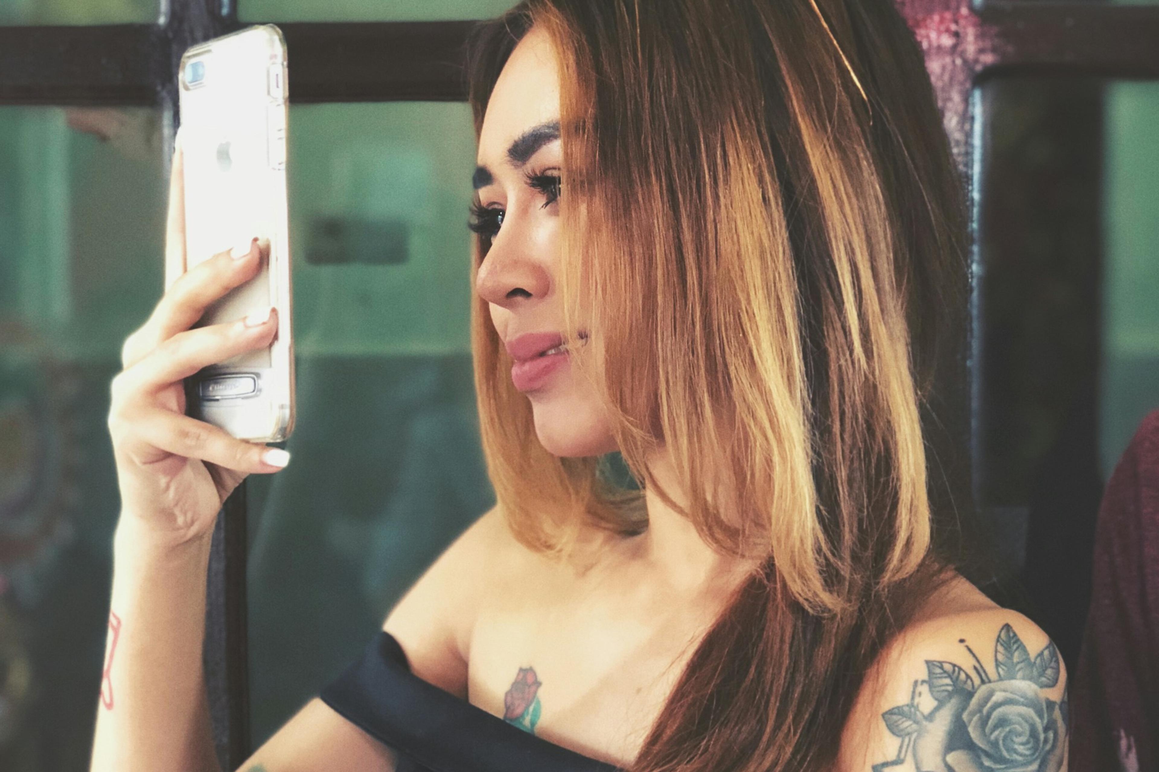 Photo of a woman with tattoos taking a selfie on a smartphone wearing an off-shoulder dress in front of glass doors.