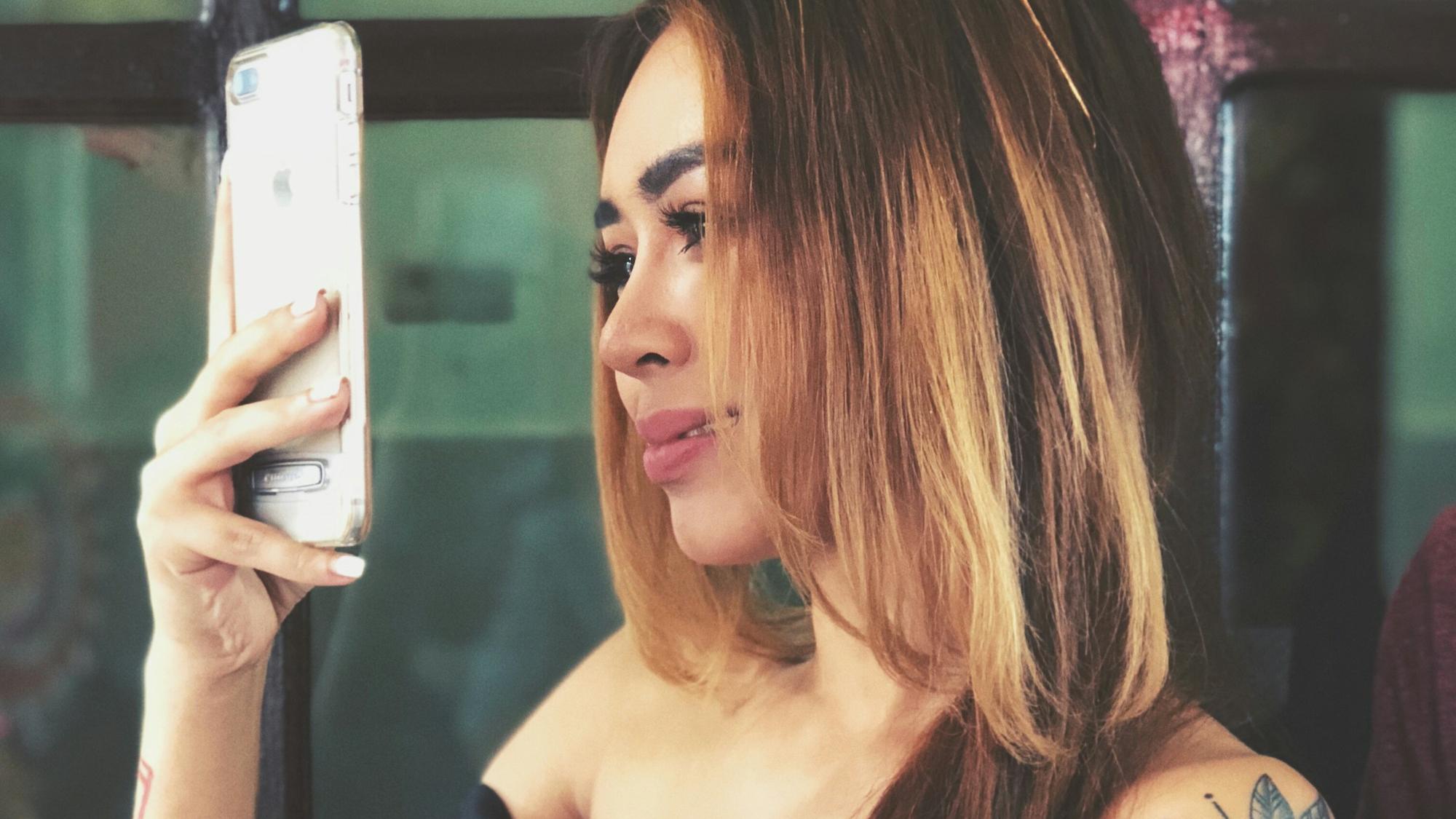Photo of a woman with tattoos taking a selfie on a smartphone wearing an off-shoulder dress in front of glass doors.
