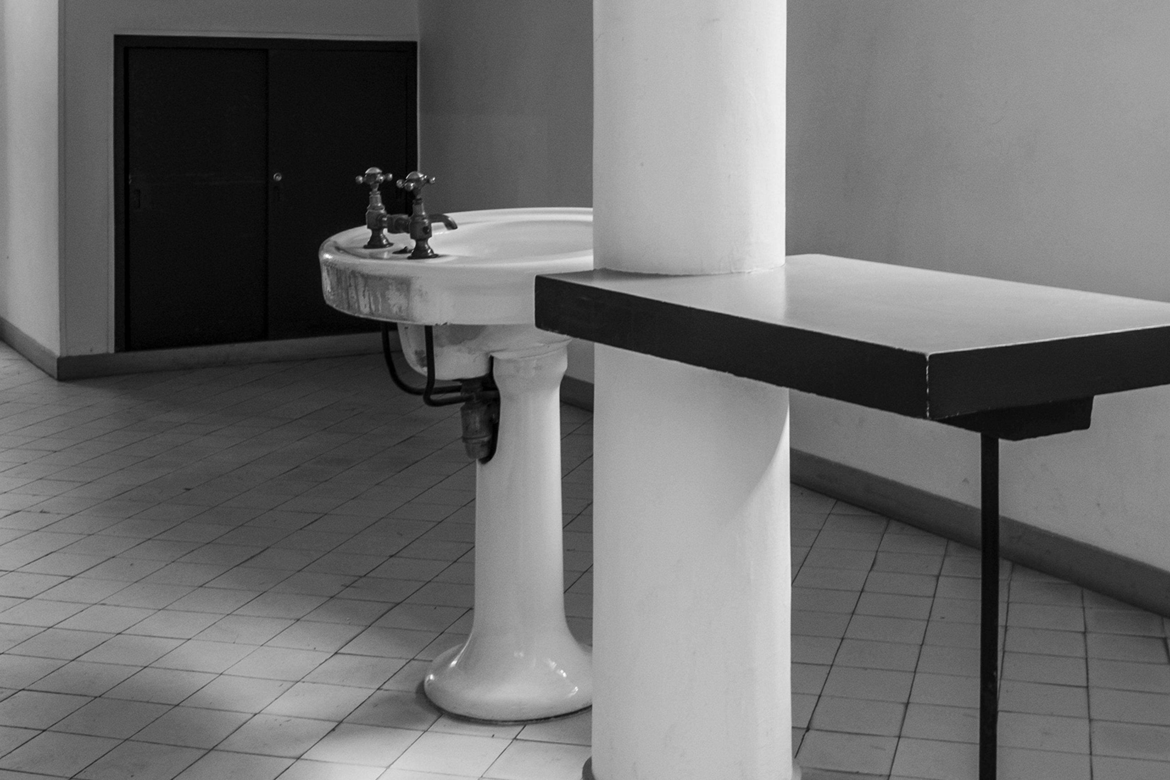 Black and white photo of a bathroom with a pedestal sink, two taps and a geometric counter, on a tiled floor.