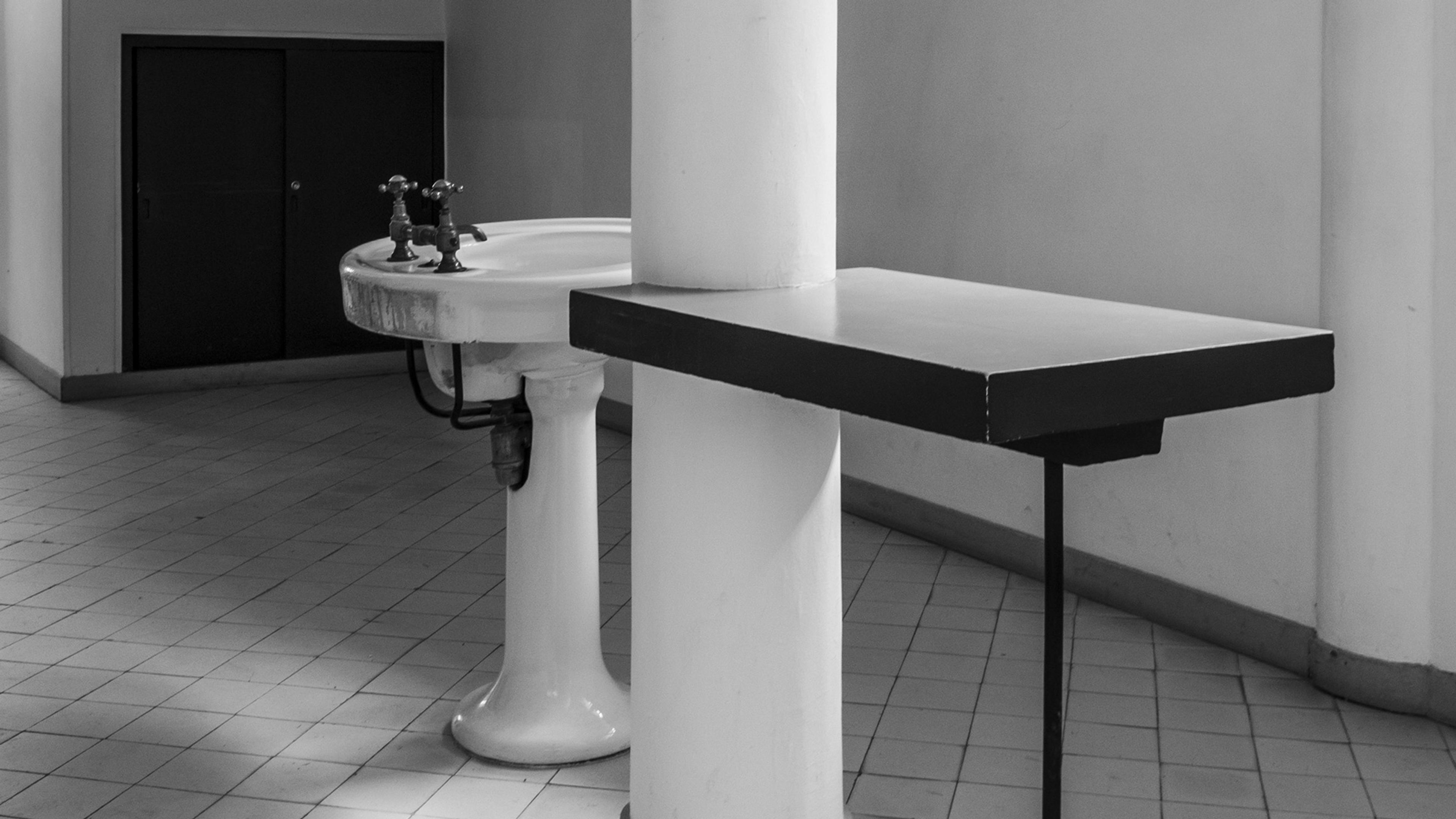 Black and white photo of a bathroom with a pedestal sink, two taps and a geometric counter, on a tiled floor.