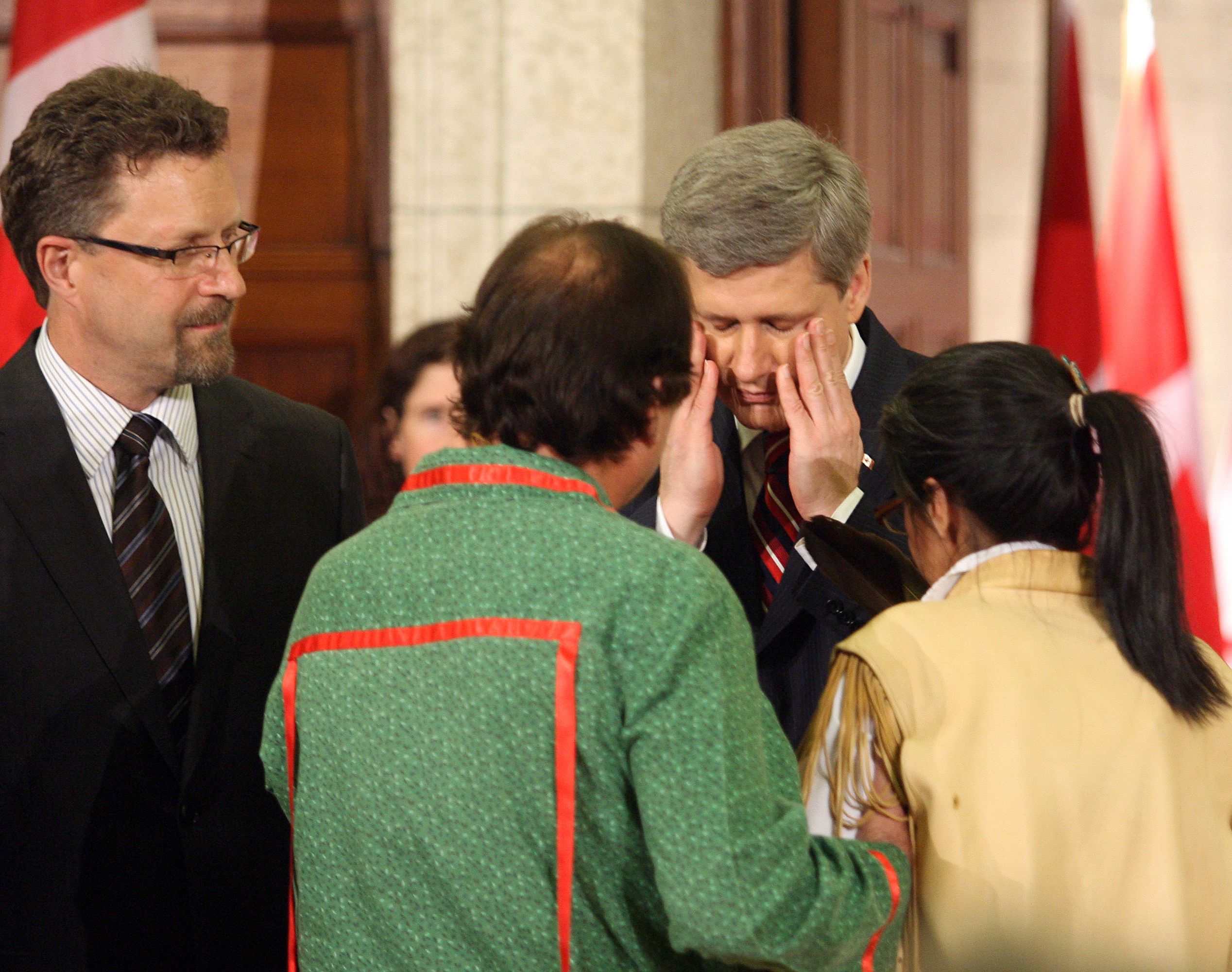 A suited man interacting with people in traditional clothing, flags in the background.