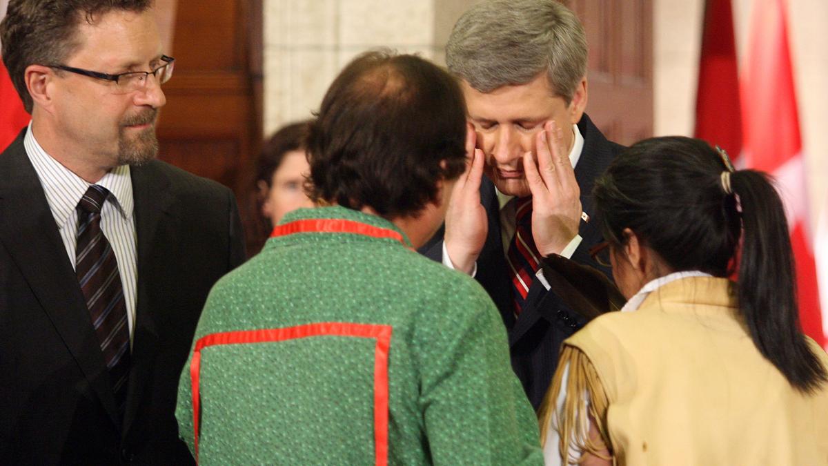 A suited man interacting with people in traditional clothing, flags in the background.