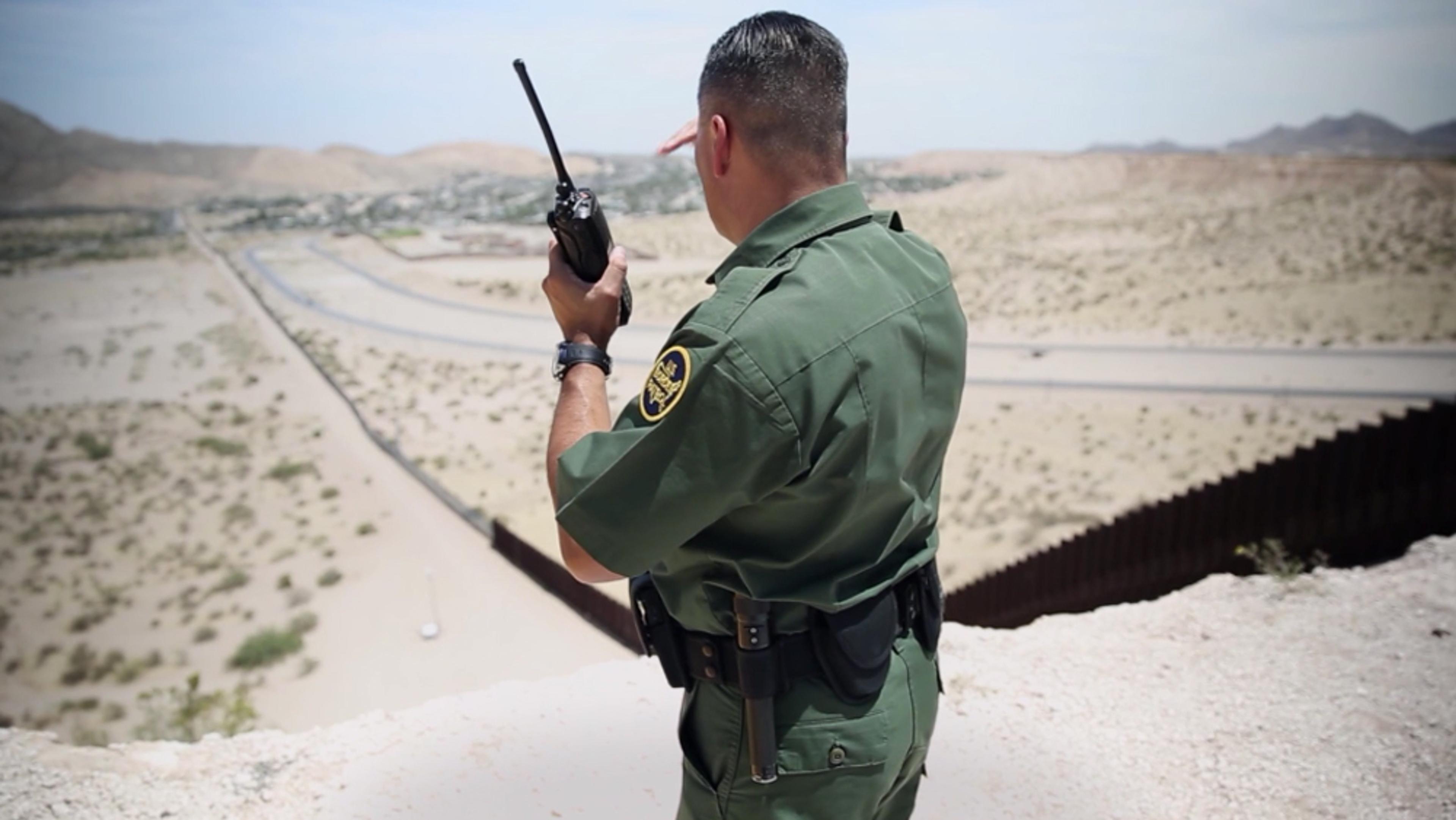 A border patrol agent in green uniform holding a radio on a desert overlook near a border fence in a mountainous area.