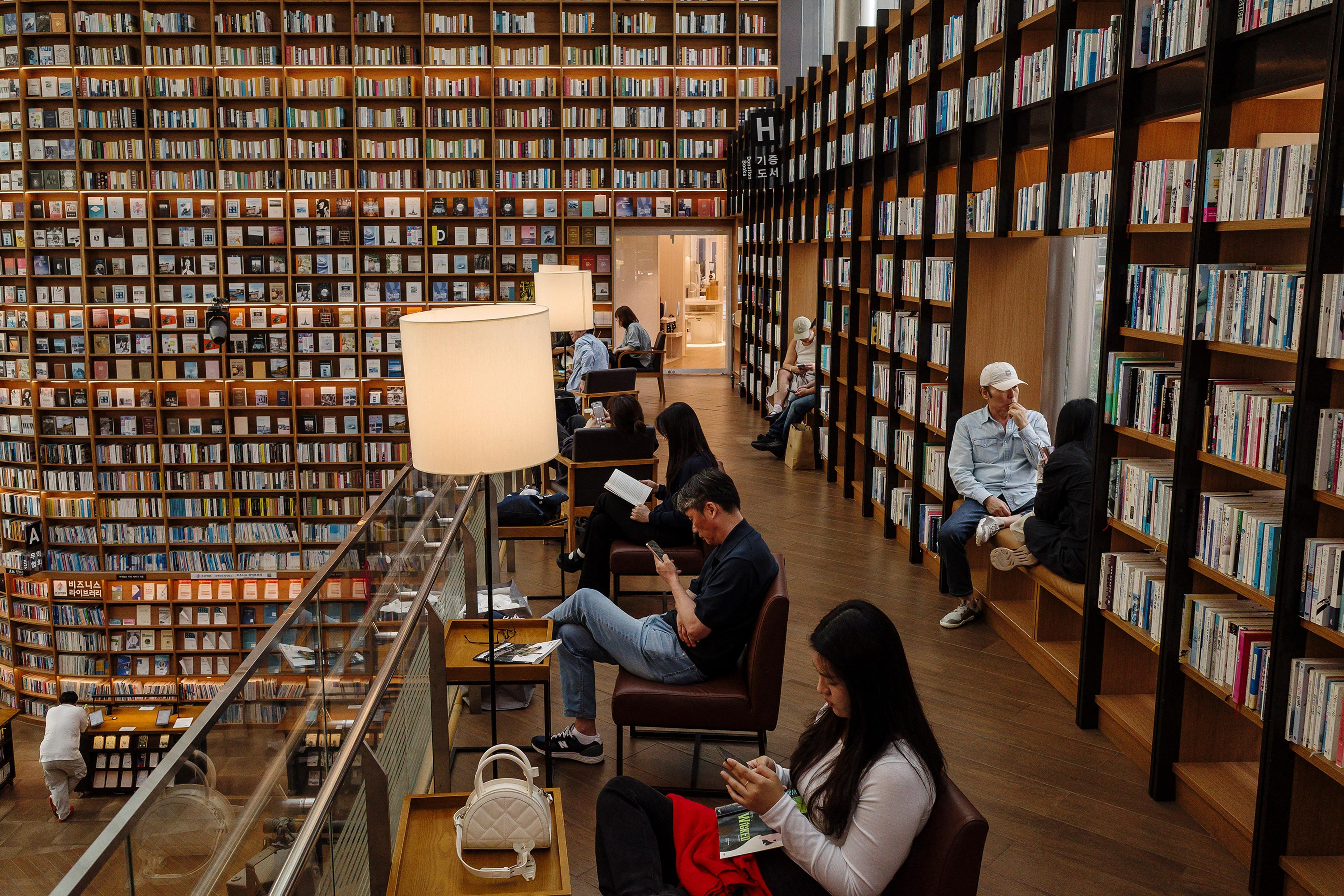 A modern library with tall bookshelves and people reading or using devices in a cosy, well-lit atmosphere.