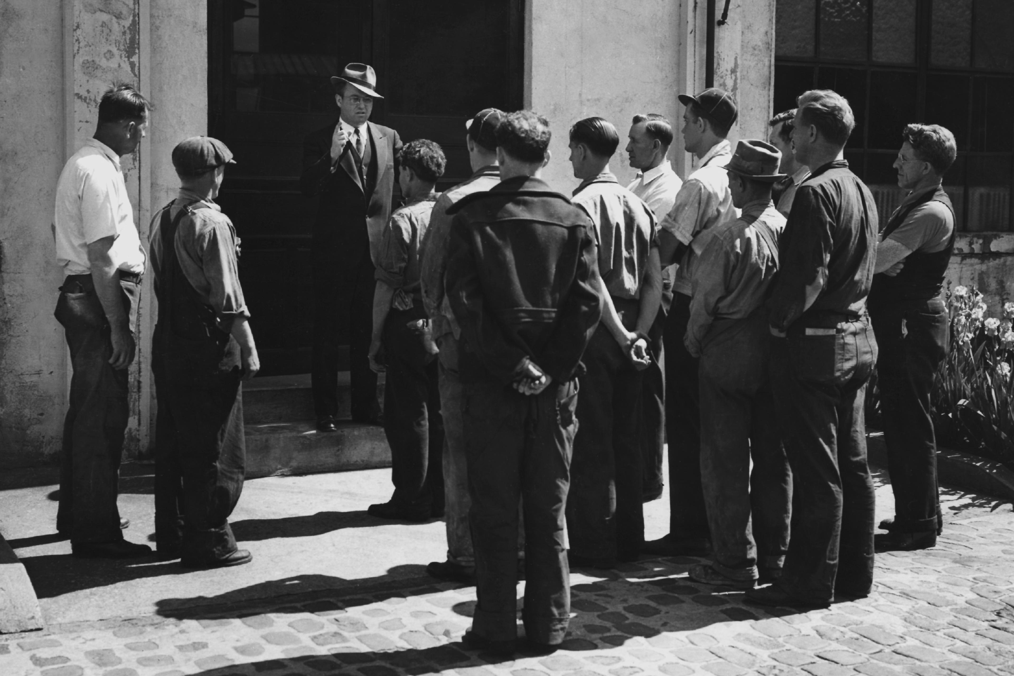 Black and white photo of a man in a suit addressing a group of workers outside a building.
