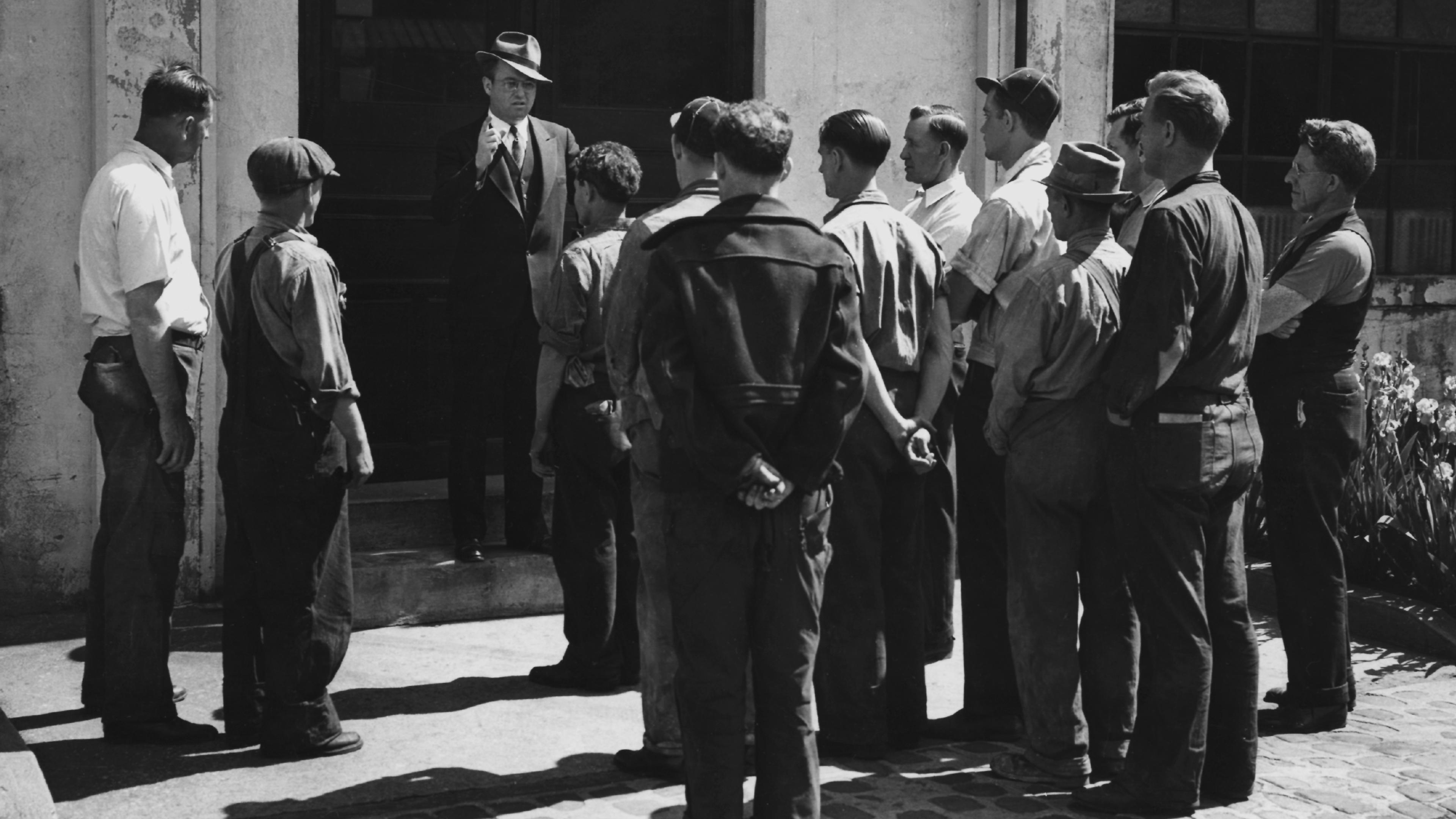 Black and white photo of a man in a suit addressing a group of workers outside a building.