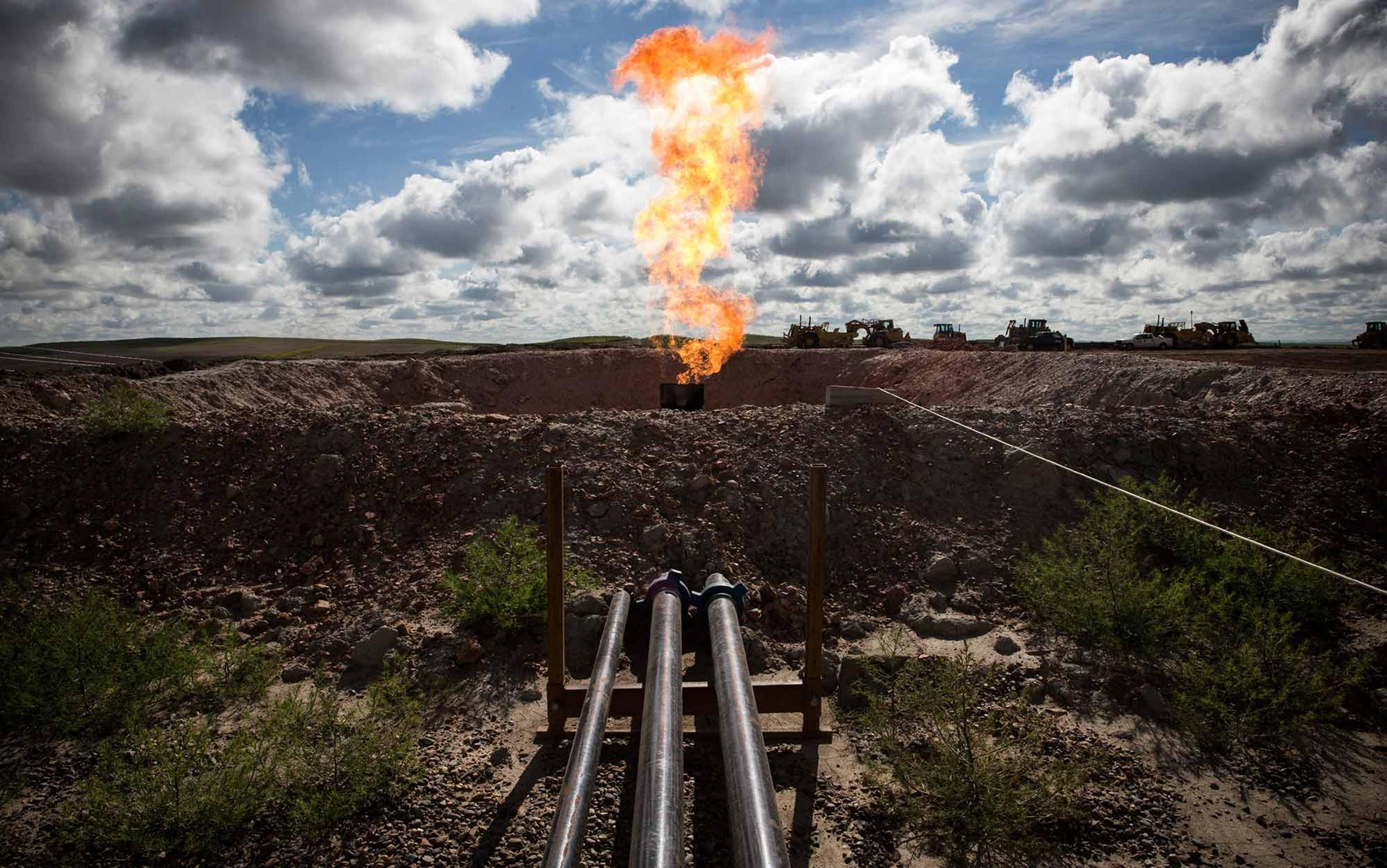 Three pipelines leading to a gas flare in a rocky, open field under a partly cloudy sky with construction vehicles in the background.