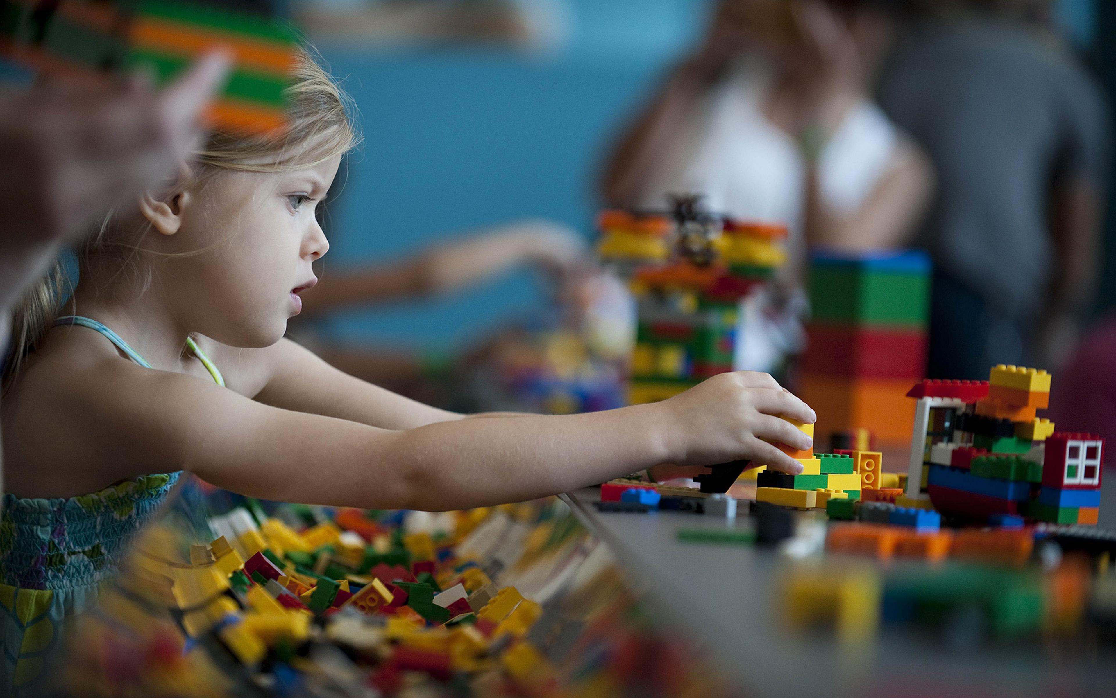 A child playing with Lego bricks, focused on building a colourful structure, with other children blurred in the background.