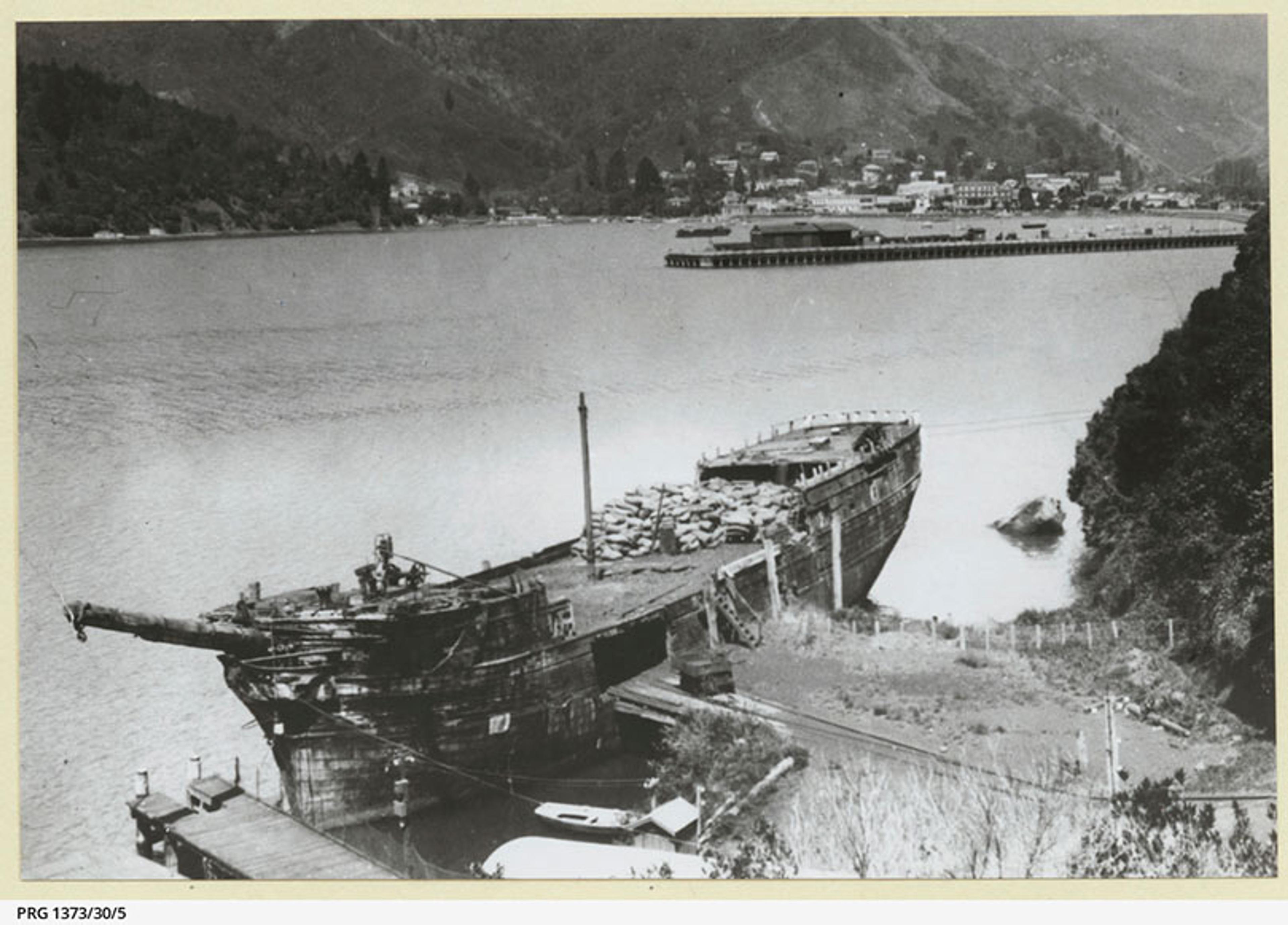 A black and white photo of an old shipwreck on the shore with a town and dock visible across the water, surrounded by mountains.