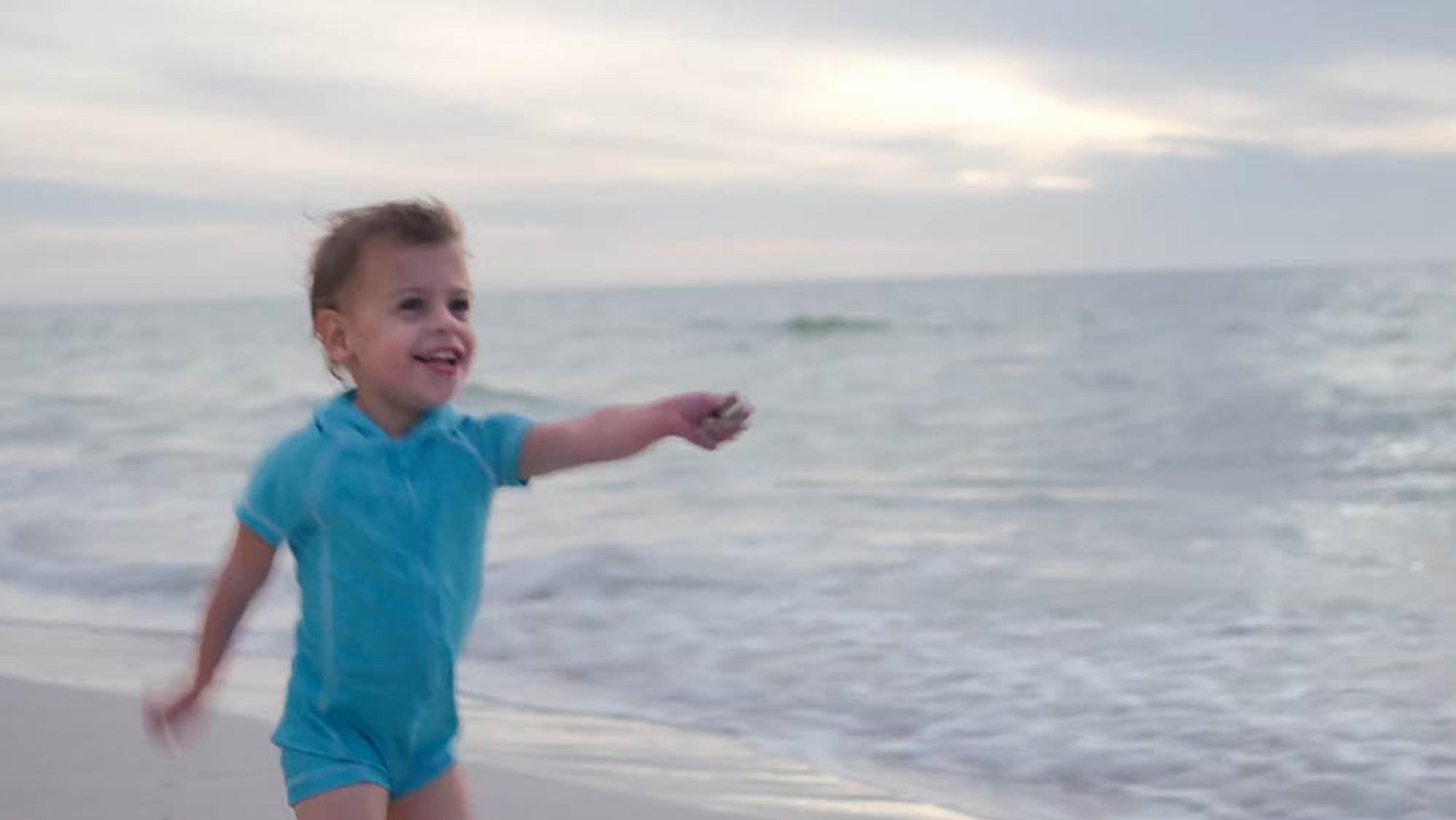 A young child in a blue outfit, smiling and running along a beach by the sea on a cloudy day.