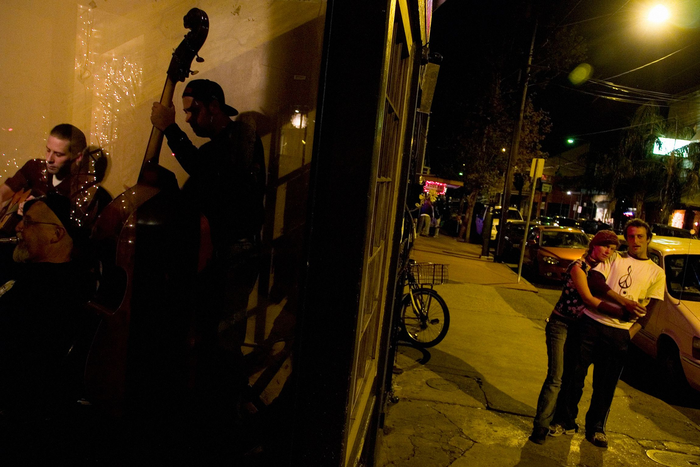 A nighttime street scene with musicians performing indoors on the left and a couple embracing outside on the right.