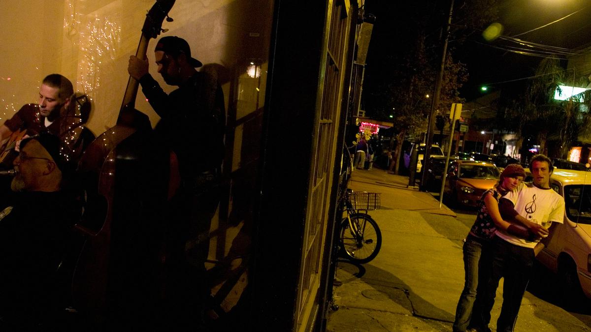 A nighttime street scene with musicians performing indoors on the left and a couple embracing outside on the right.