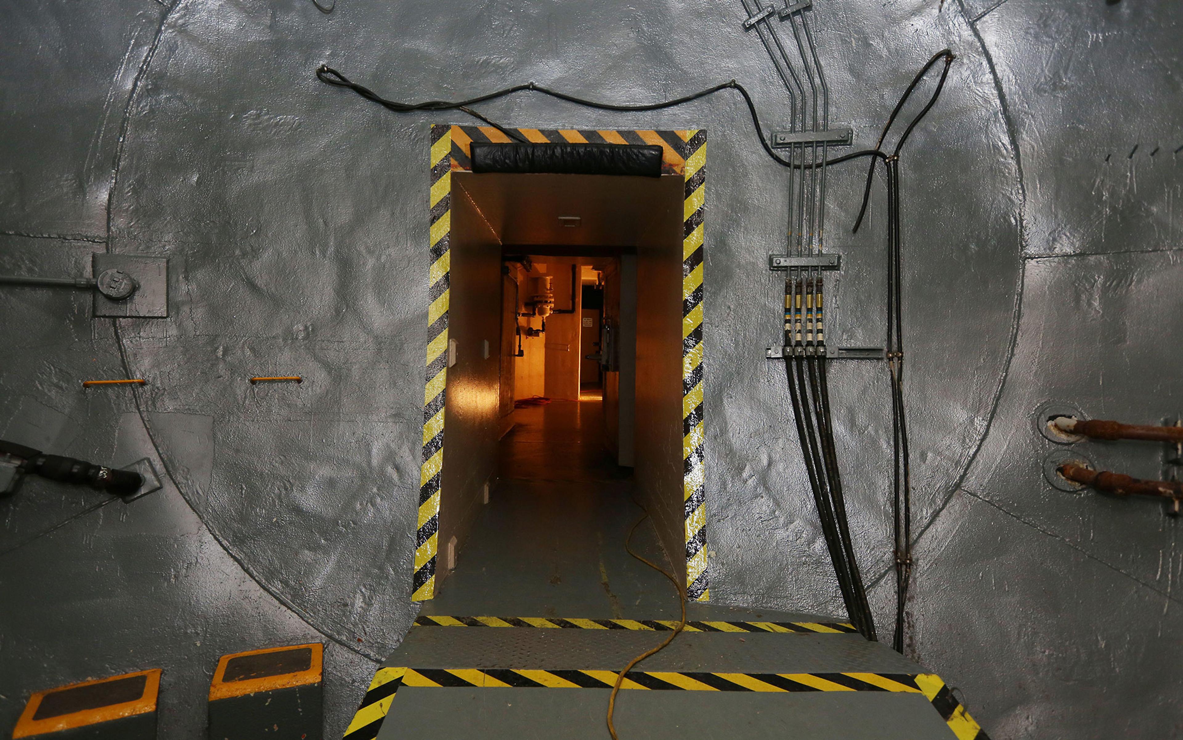 A doorway within a metallic-like surface surrounded by hazard tape leads to a low-lit tunnel
