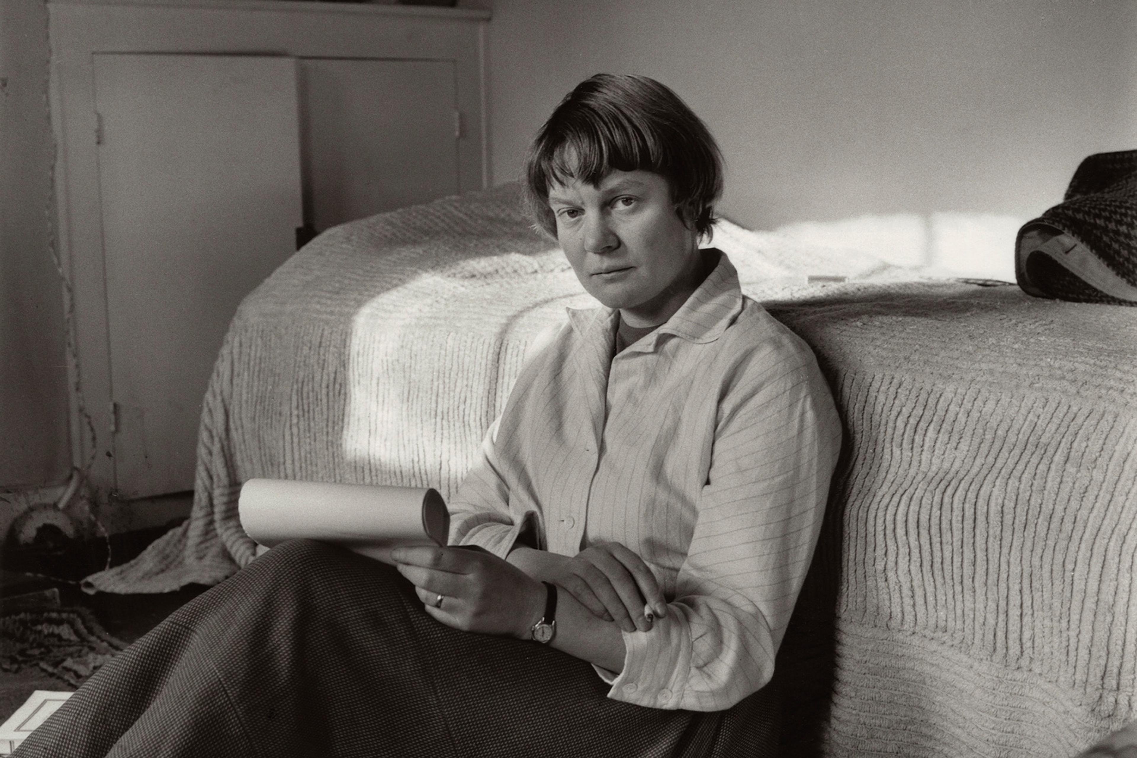 Black and white photo of a person sitting on a floor by a bed holding papers, with books and papers scattered around.