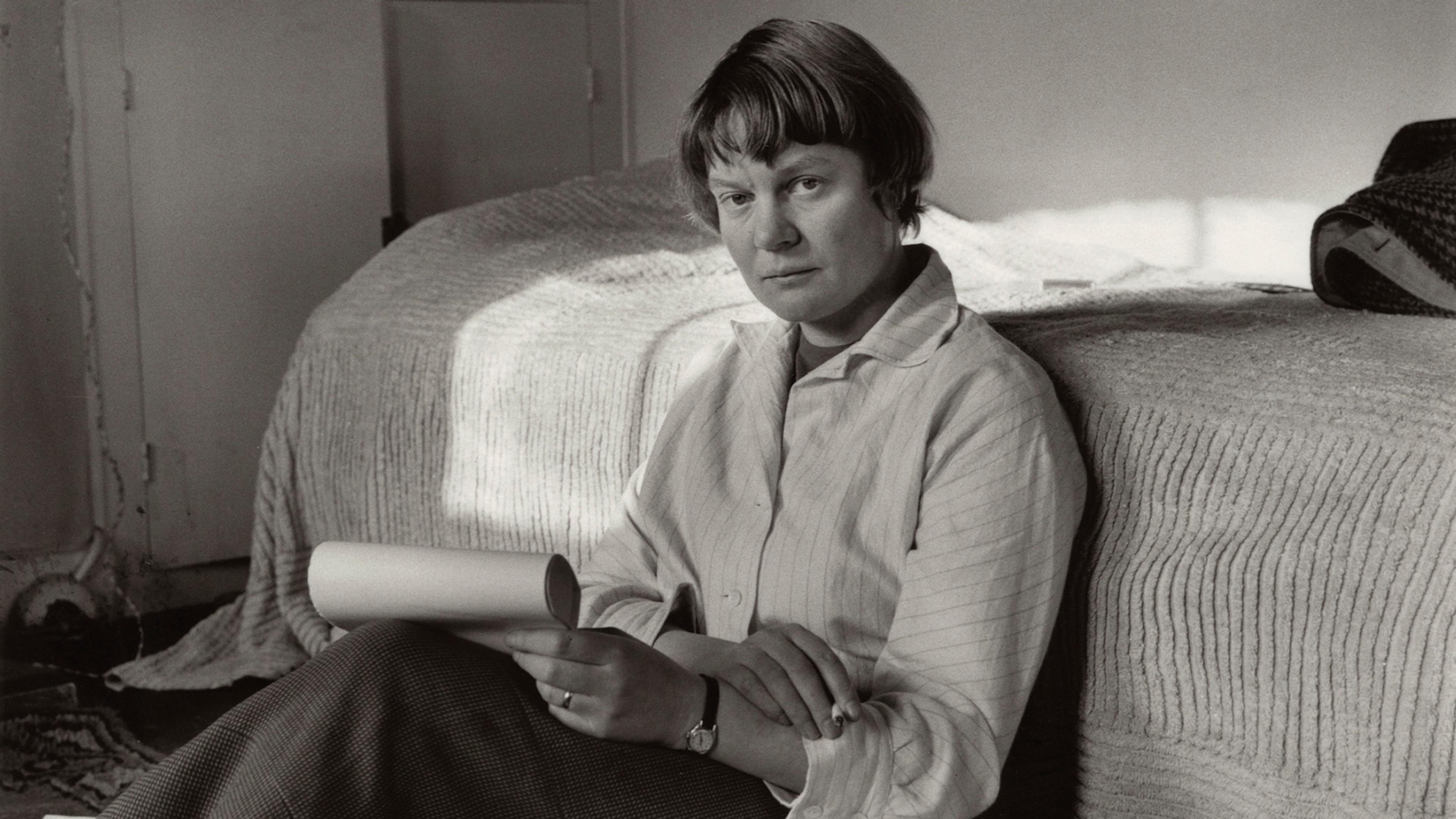 Black and white photo of a person sitting on a floor by a bed holding papers, with books and papers scattered around.