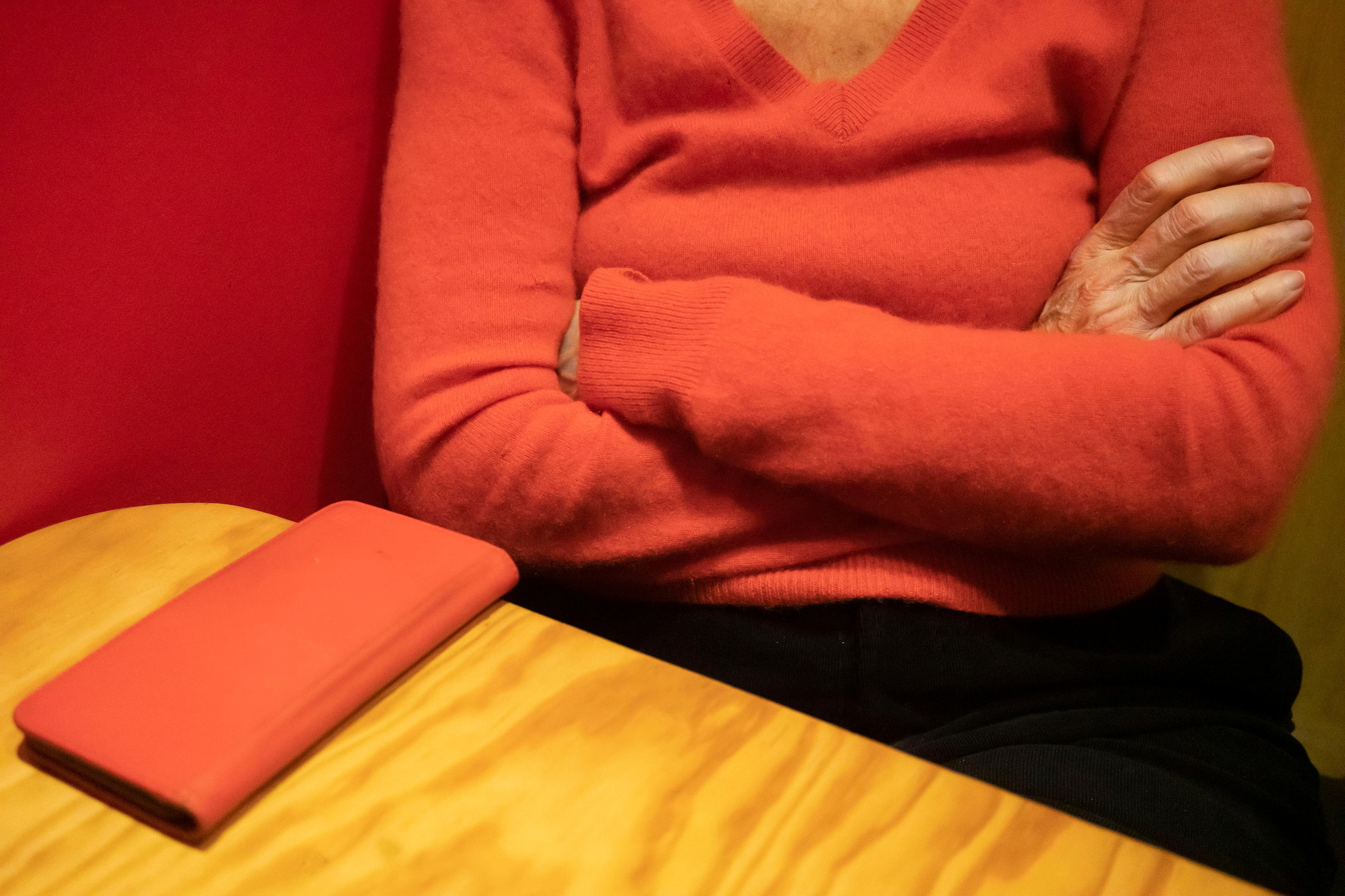 A person in a red jumper with folded arms, a closed red phone case is placed on a wooden table in front of the person.