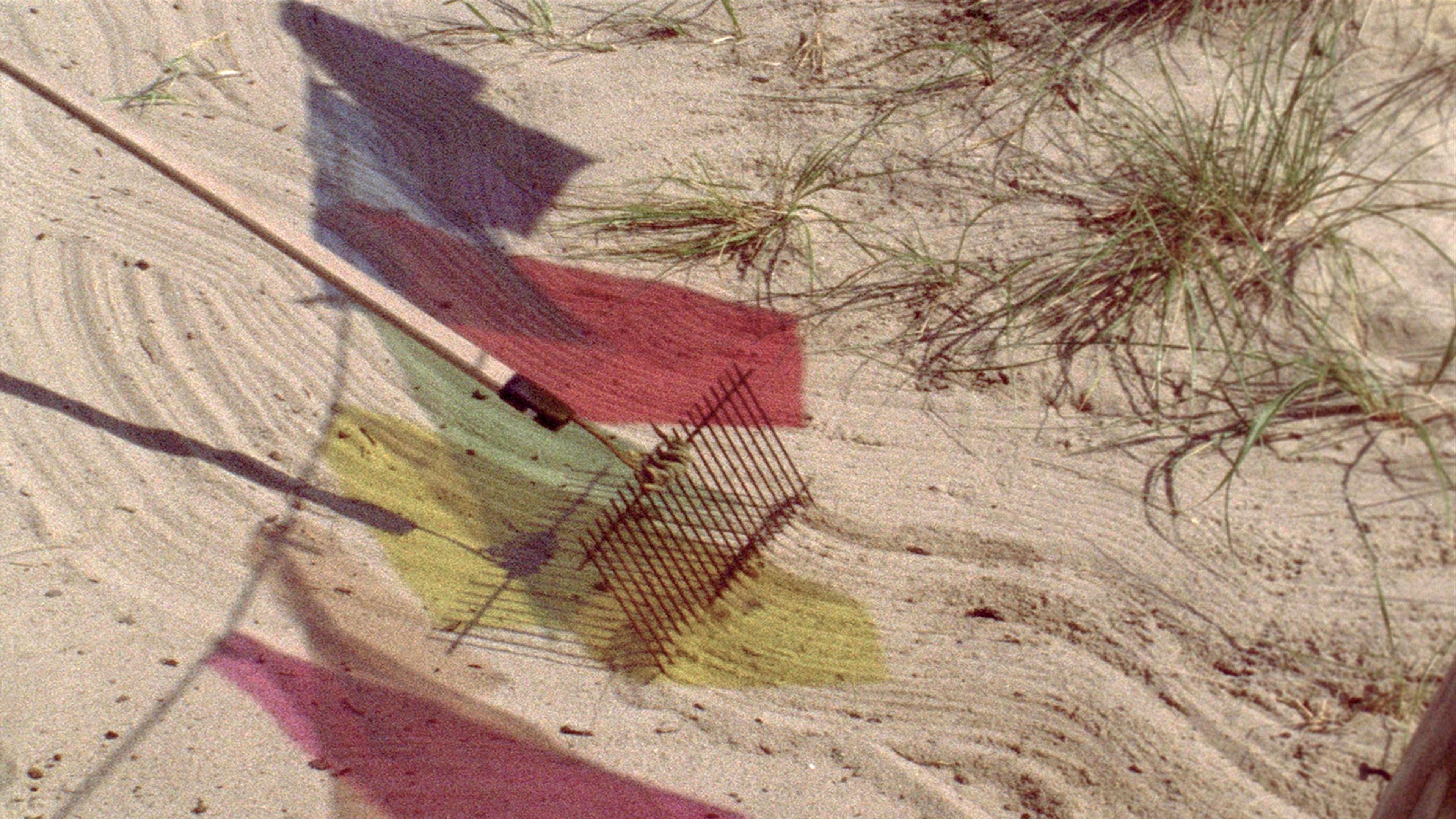 A rake with a wooden handle rakes through sand dunes, its trail mixing with shadows from a line of multicoloured flags.