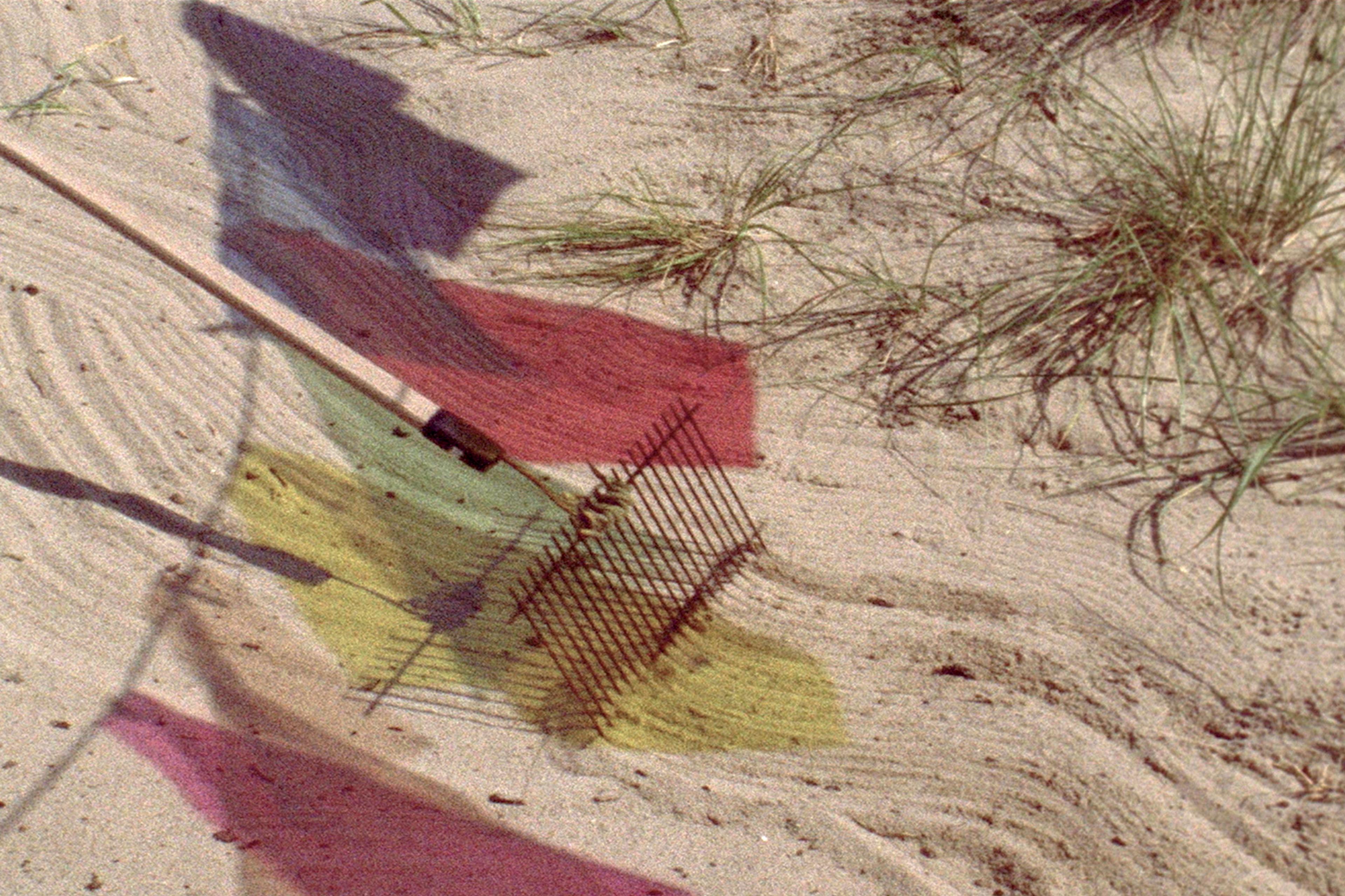 A rake with a wooden handle rakes through sand dunes, its trail mixing with shadows from a line of multicoloured flags.