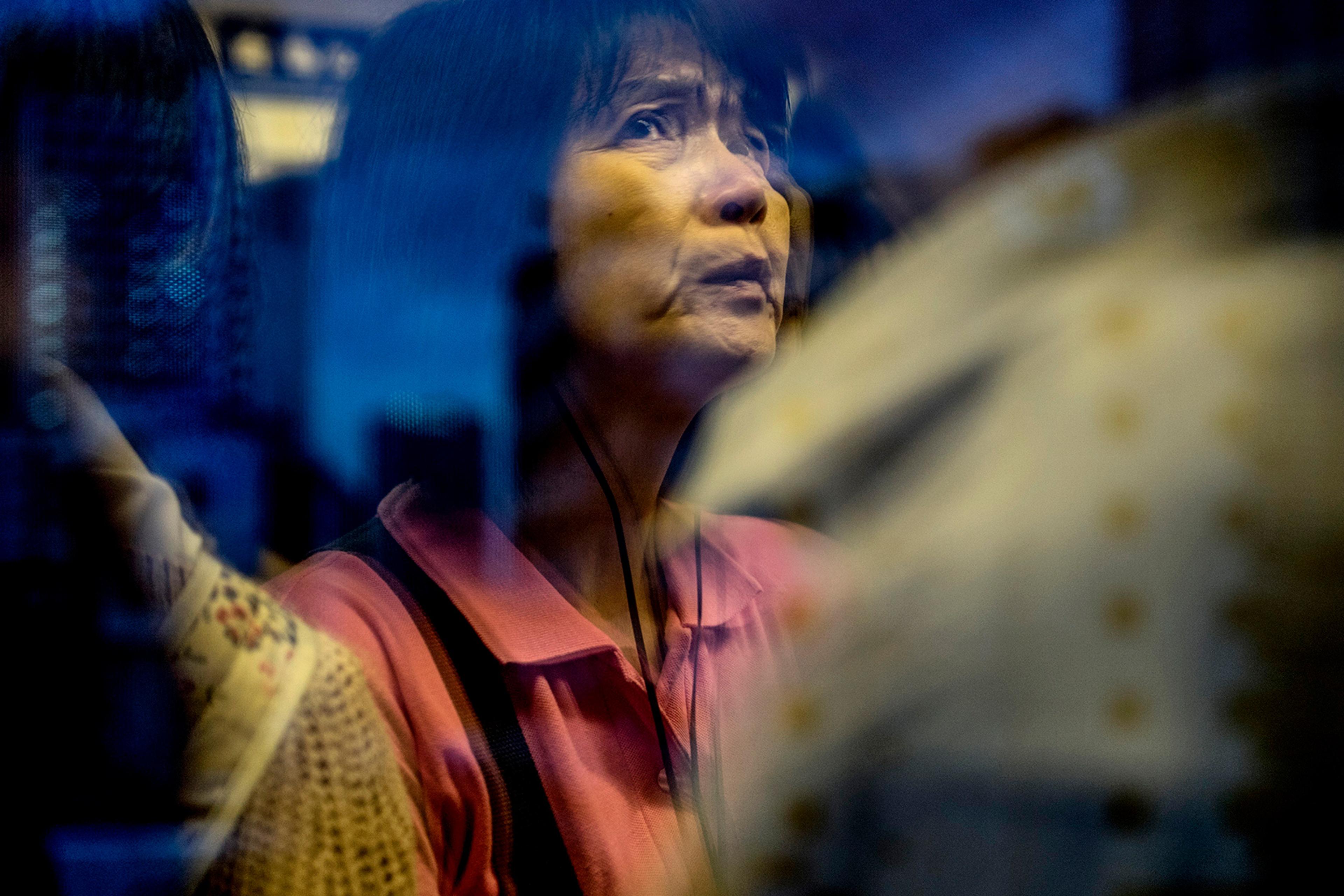 A woman in a pink shirt seen through a reflective window with a cityscape in the background, evoking a contemplative mood.