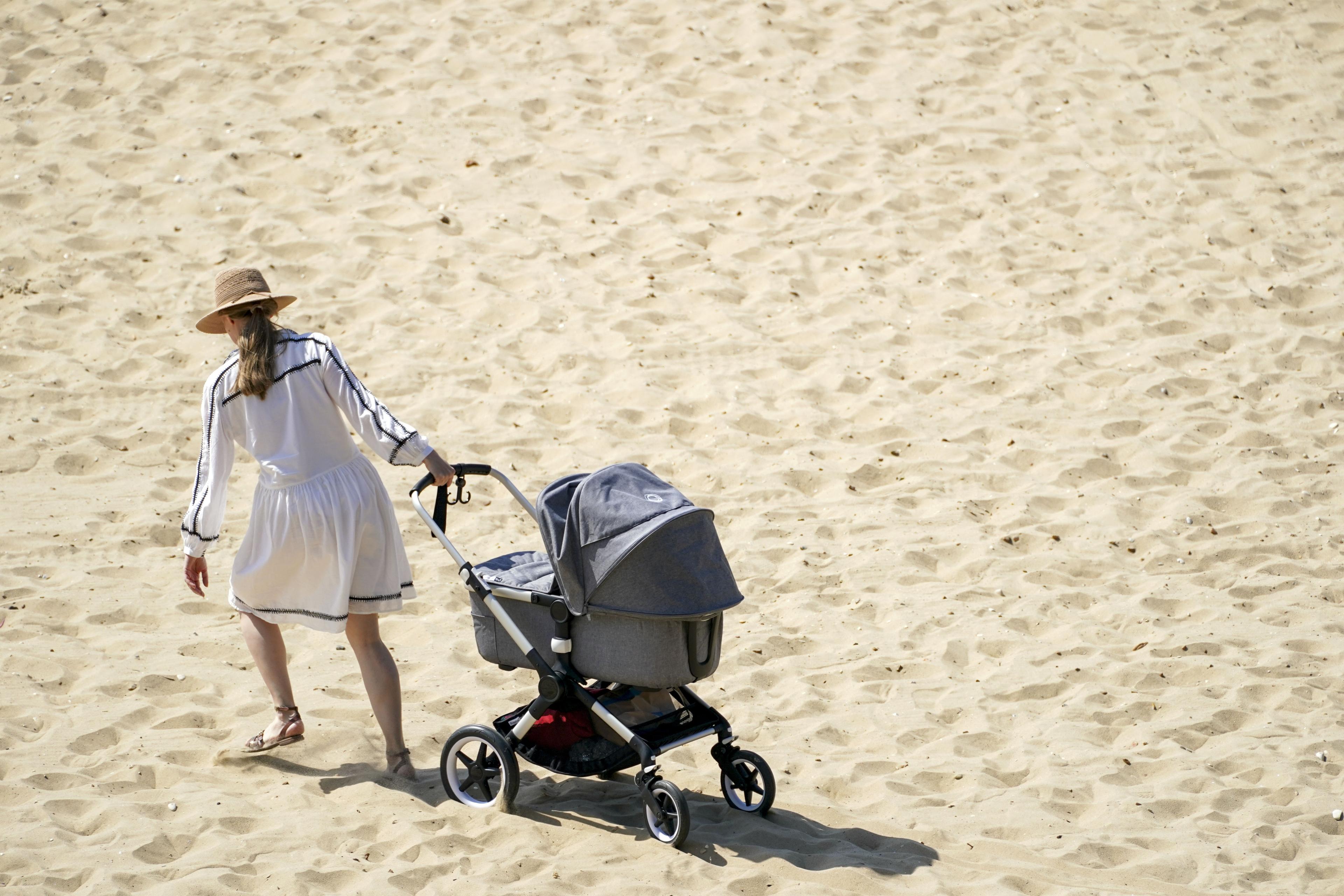 Photo of a person in a white dress and hat walking on sand pushing a grey pram.