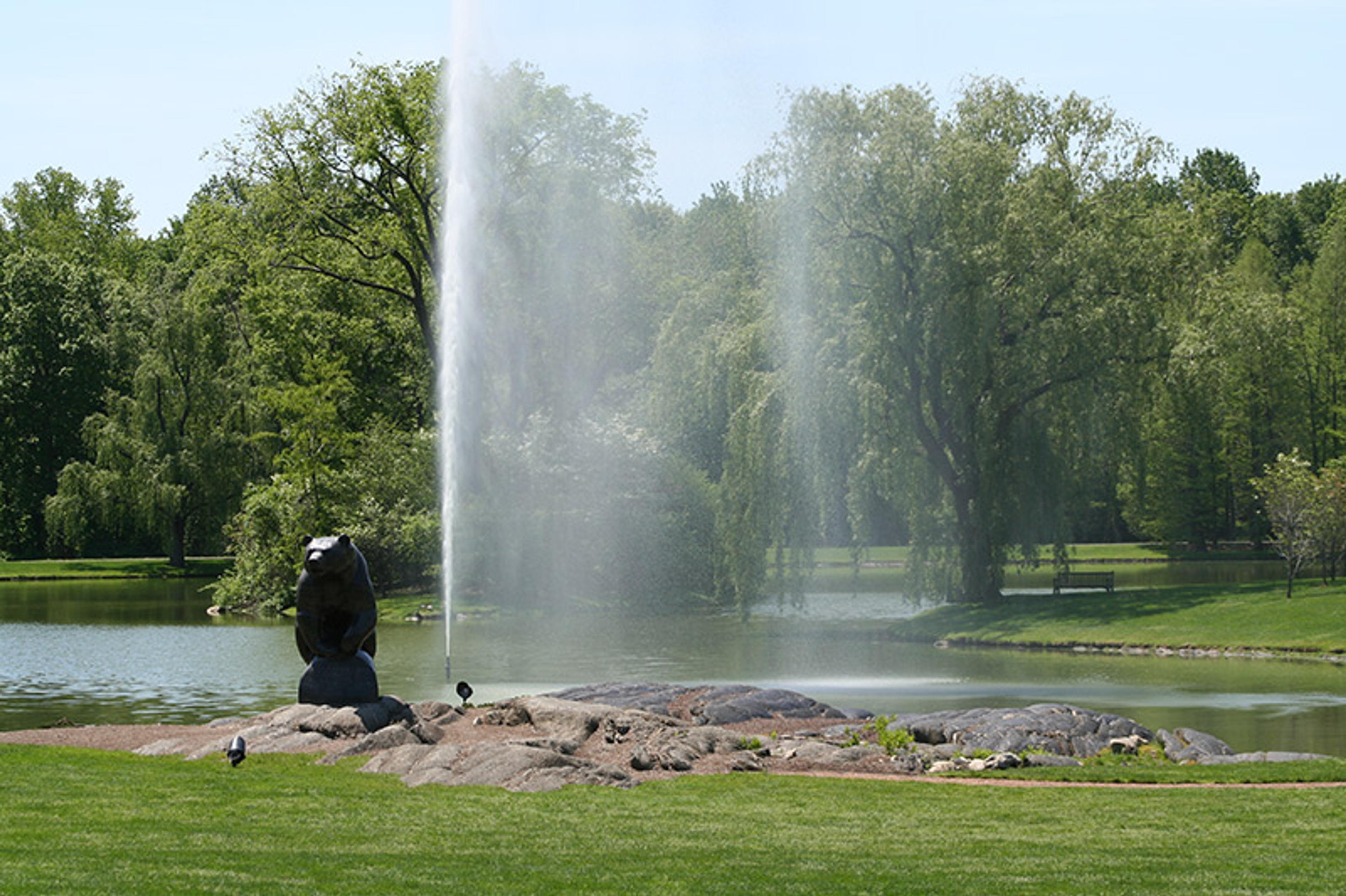 A park with a large fountain, a bear statue, a lake, green grass, and trees in the background on a clear day.