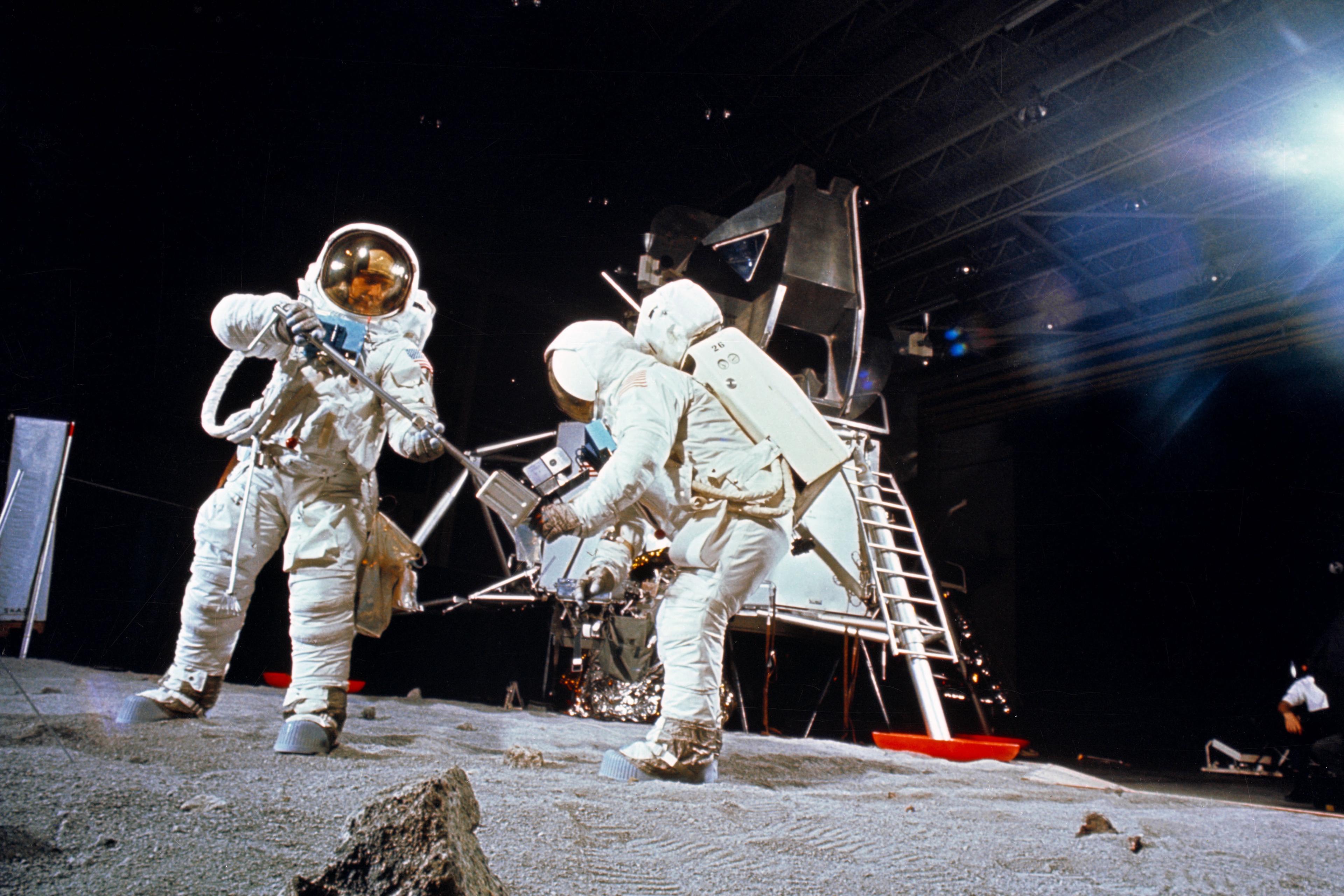 Photo of astronauts in spacesuits conducting moon surface activities near a lunar module in a studio setting.
