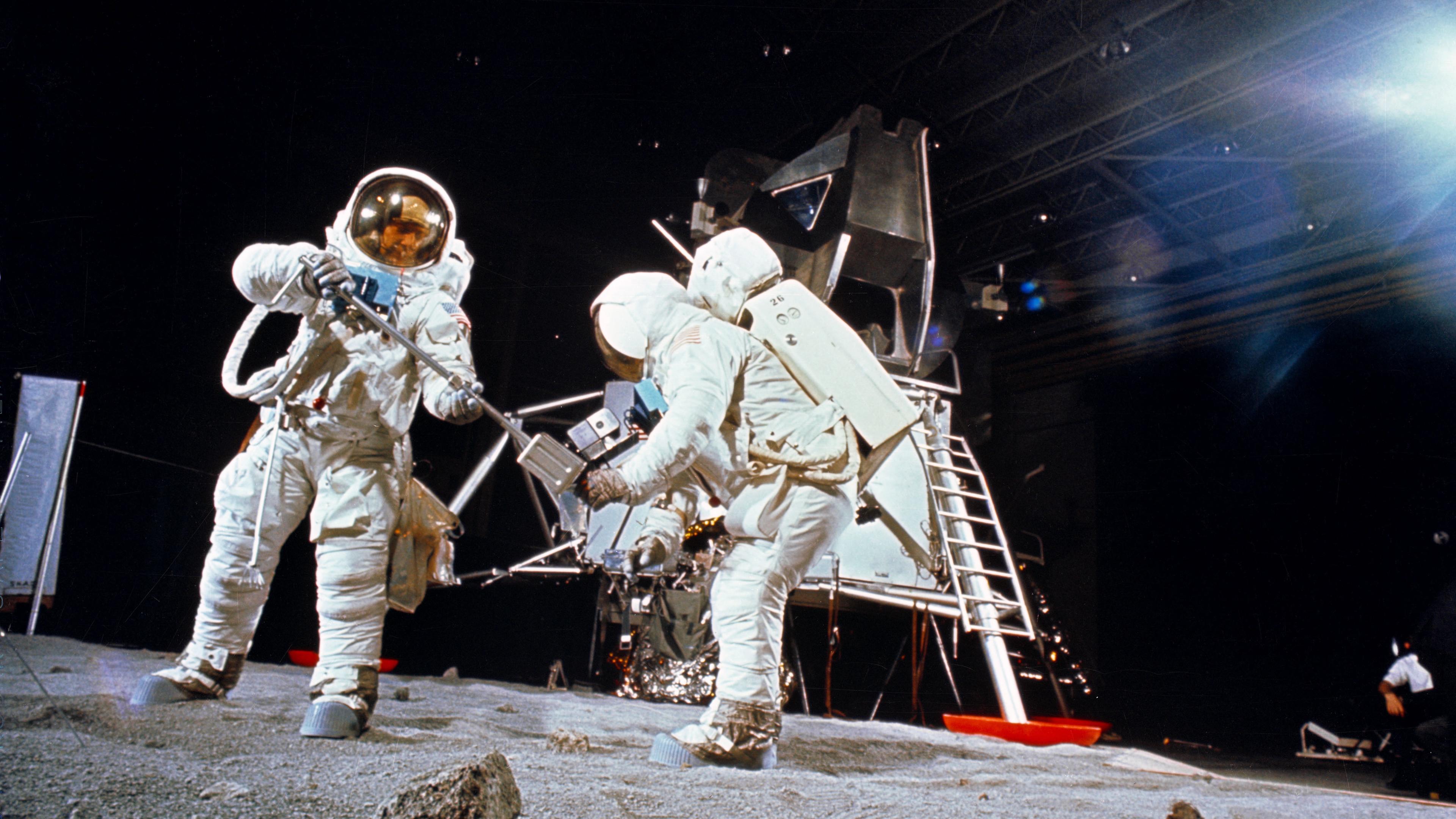 Photo of astronauts in spacesuits conducting moon surface activities near a lunar module in a studio setting.