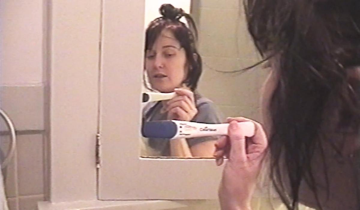 Woman holding a pregnancy test in a bathroom, looking at her reflection in the mirror with a thoughtful expression.