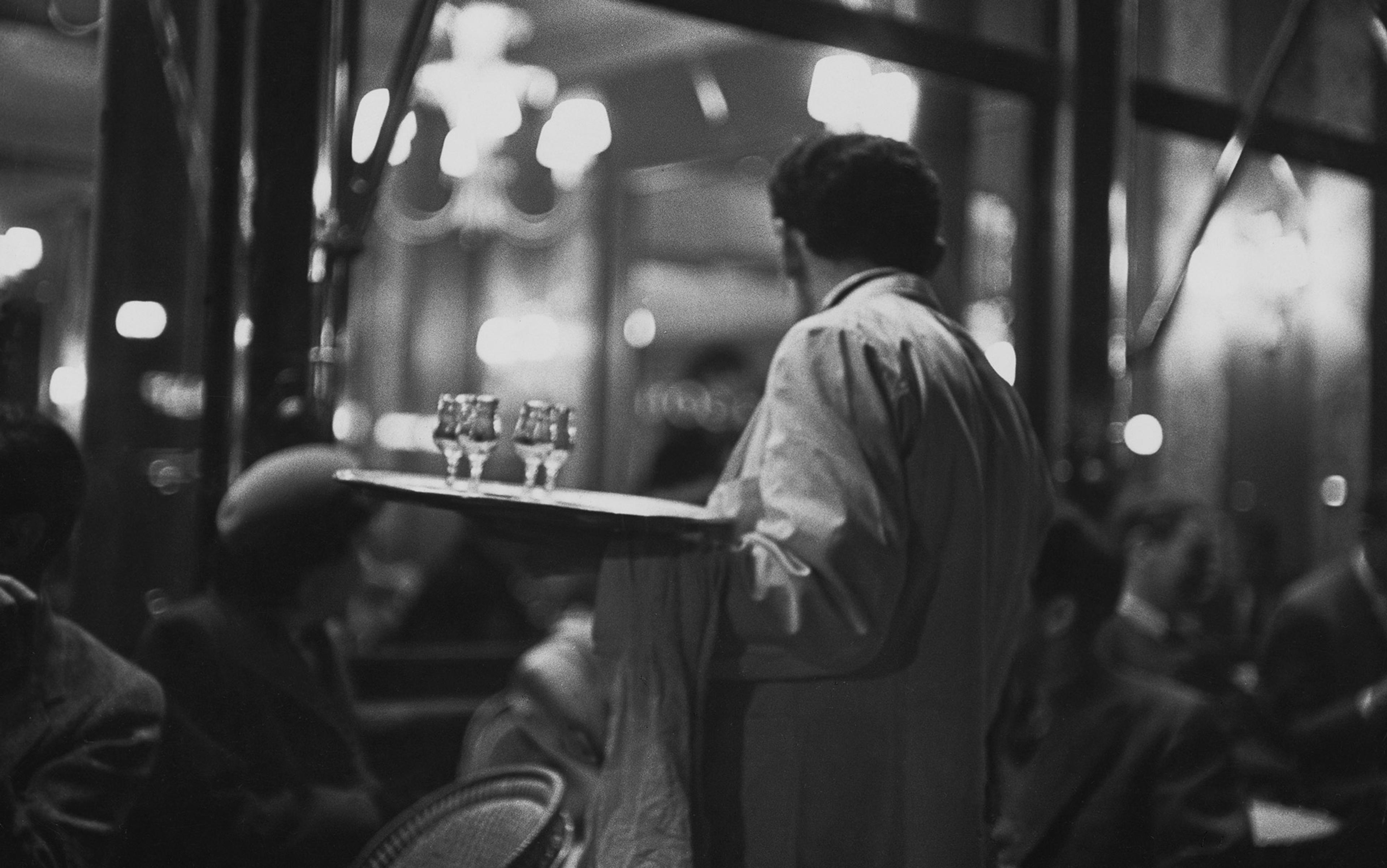 Black-and-white photo of a waiter holding a tray with glasses, serving patrons in a dimly lit, busy café or restaurant.