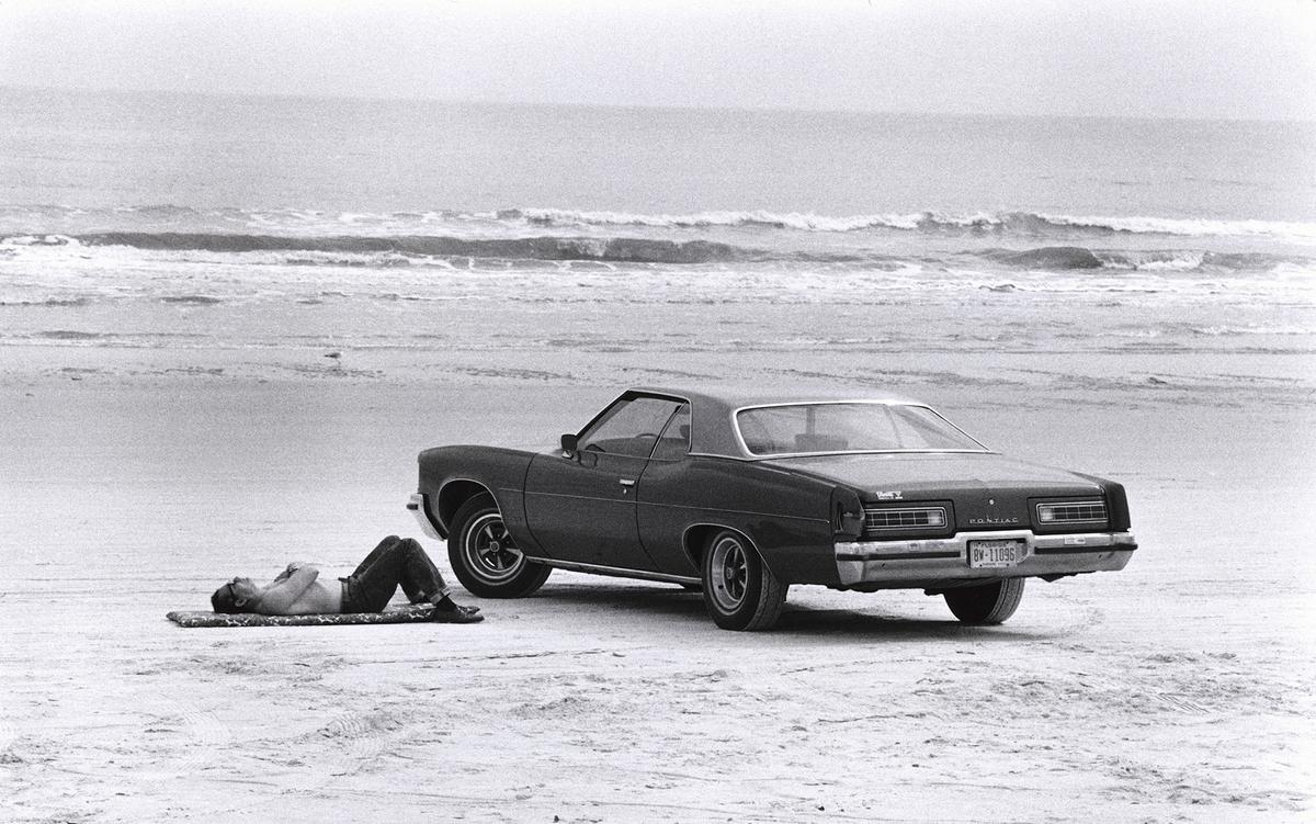 A shirtless man in jeans lies sleeping beside his 1970s era car on the sandy beach