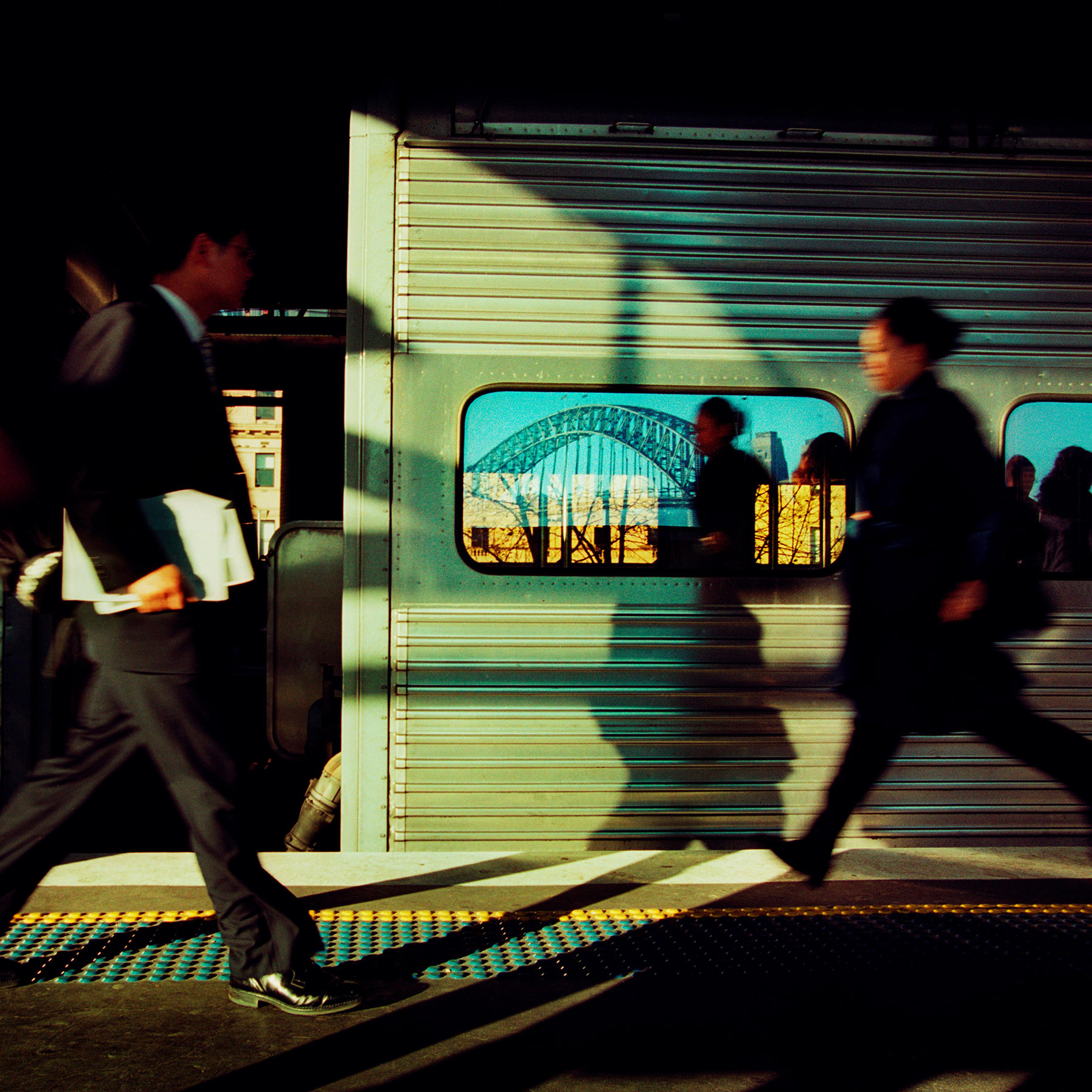 Commuters rush past a stationary commuter train. In the train window is reflected the Sydney Harbour Bridge