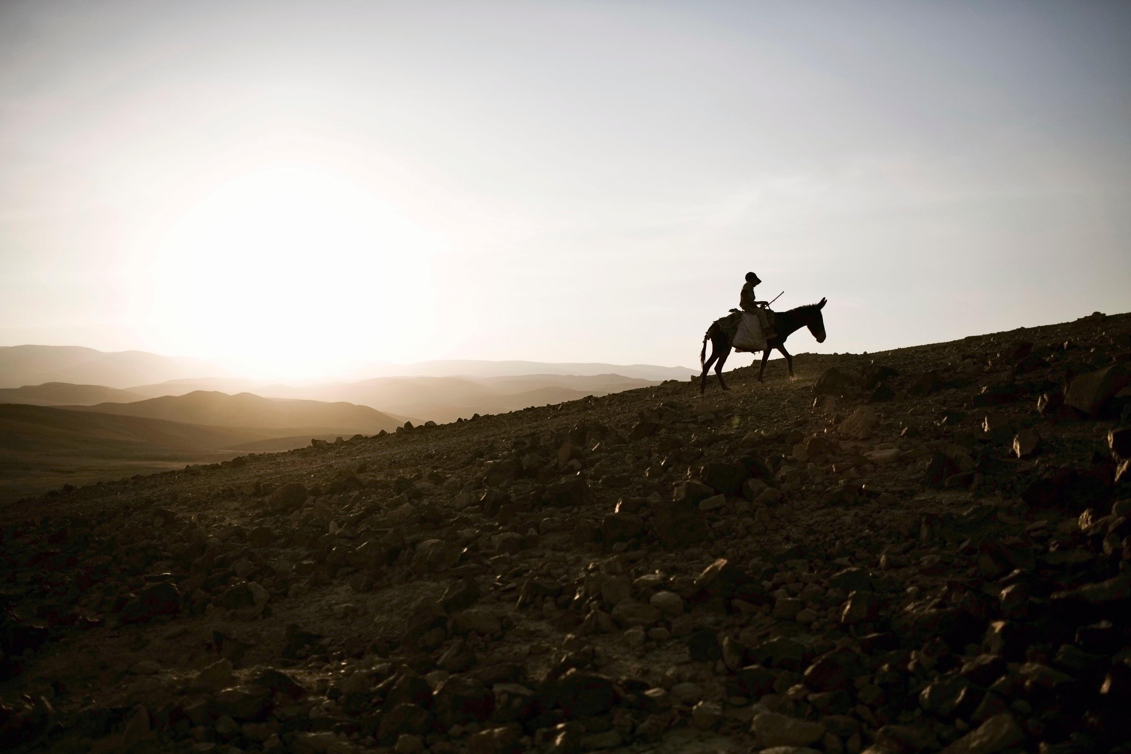 Photo of a person riding a donkey on a rocky hill at sunset with a wide, expansive sky and distant rolling hills.