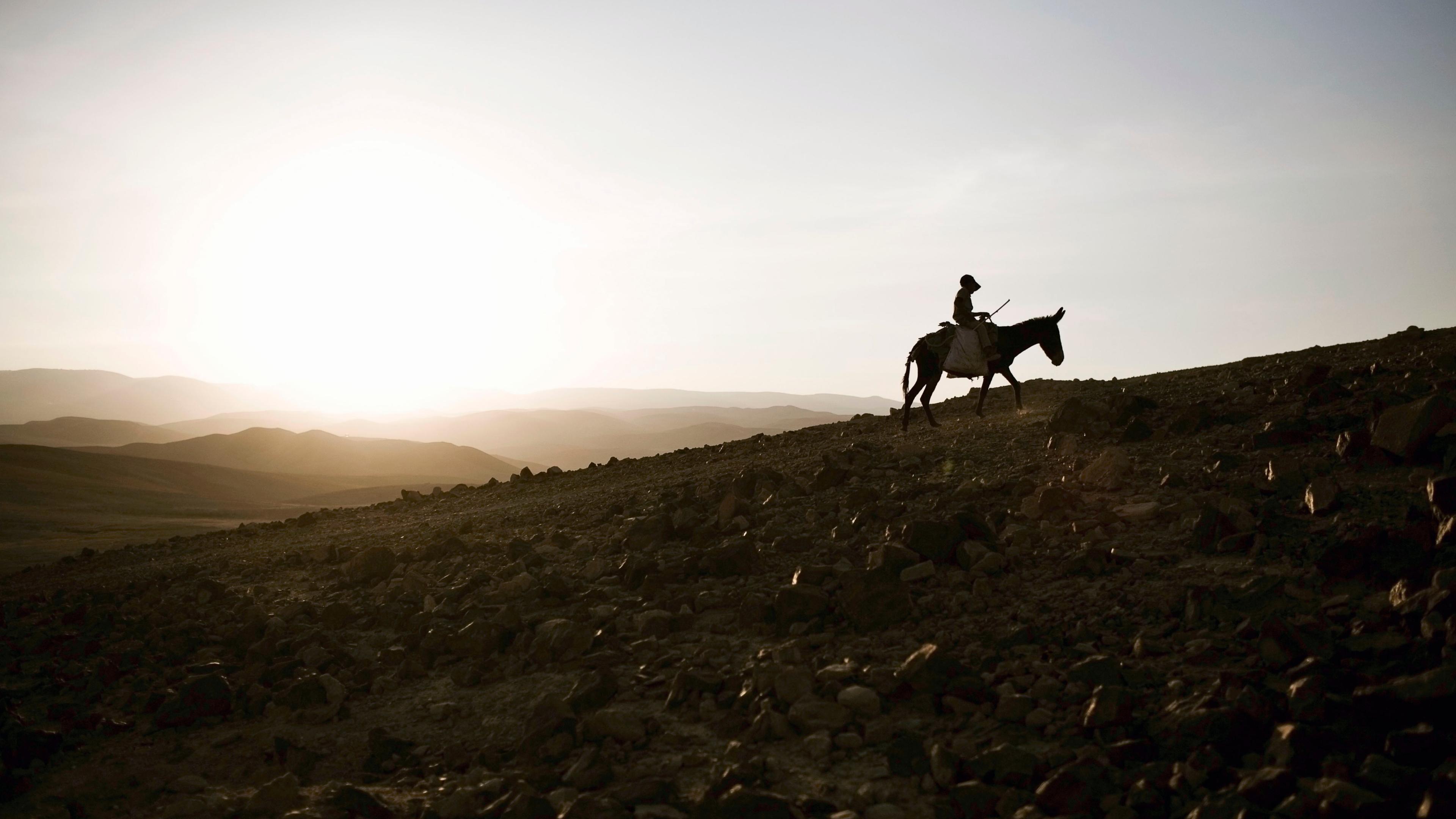 Photo of a person riding a donkey on a rocky hill at sunset with a wide, expansive sky and distant rolling hills.