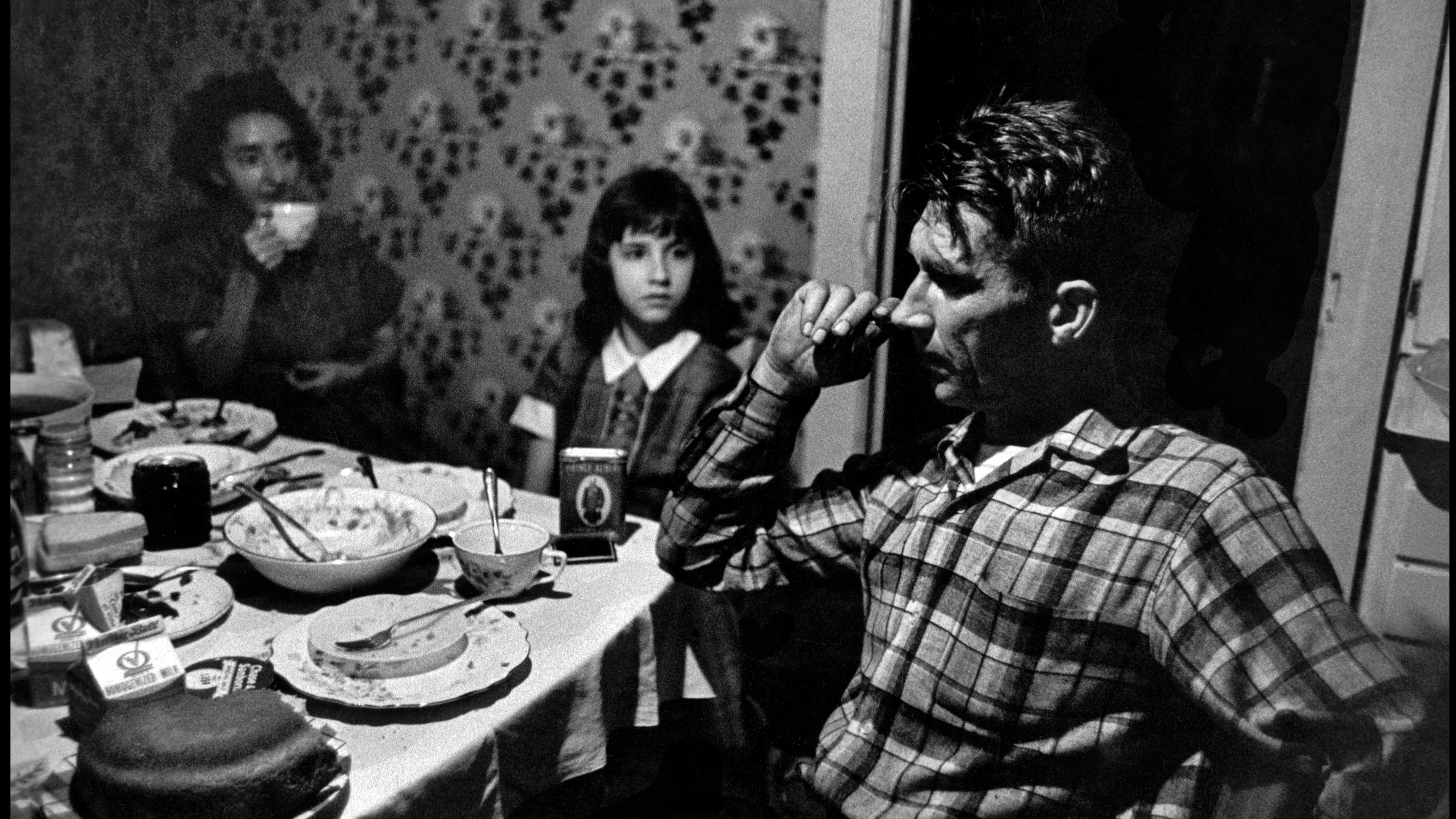 Black and white photo of a family dining, with a man in a checked shirt, a woman drinking tea and a girl looking on.