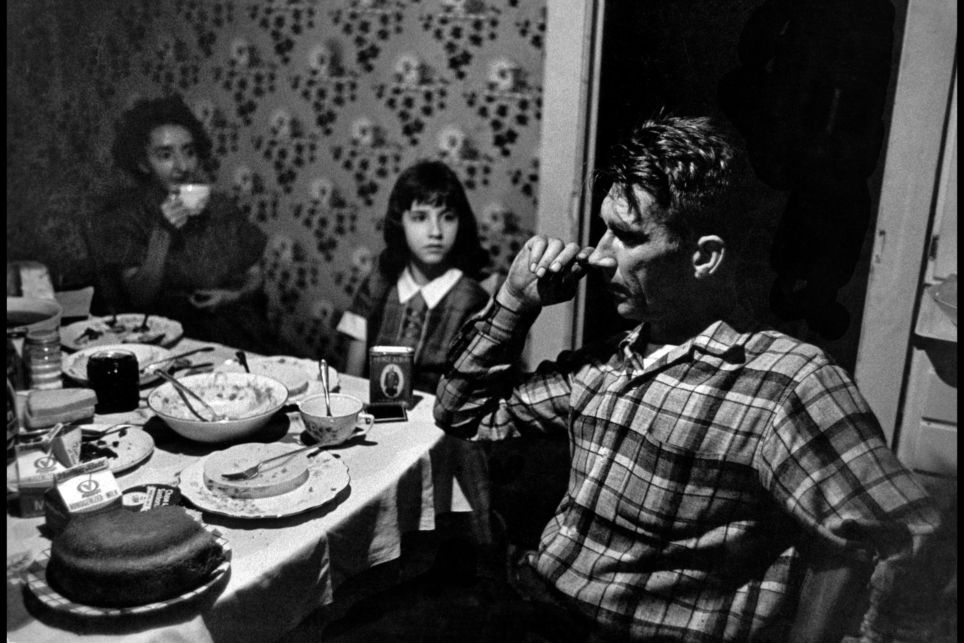 Black and white photo of a family dining, with a man in a checked shirt, a woman drinking tea and a girl looking on.