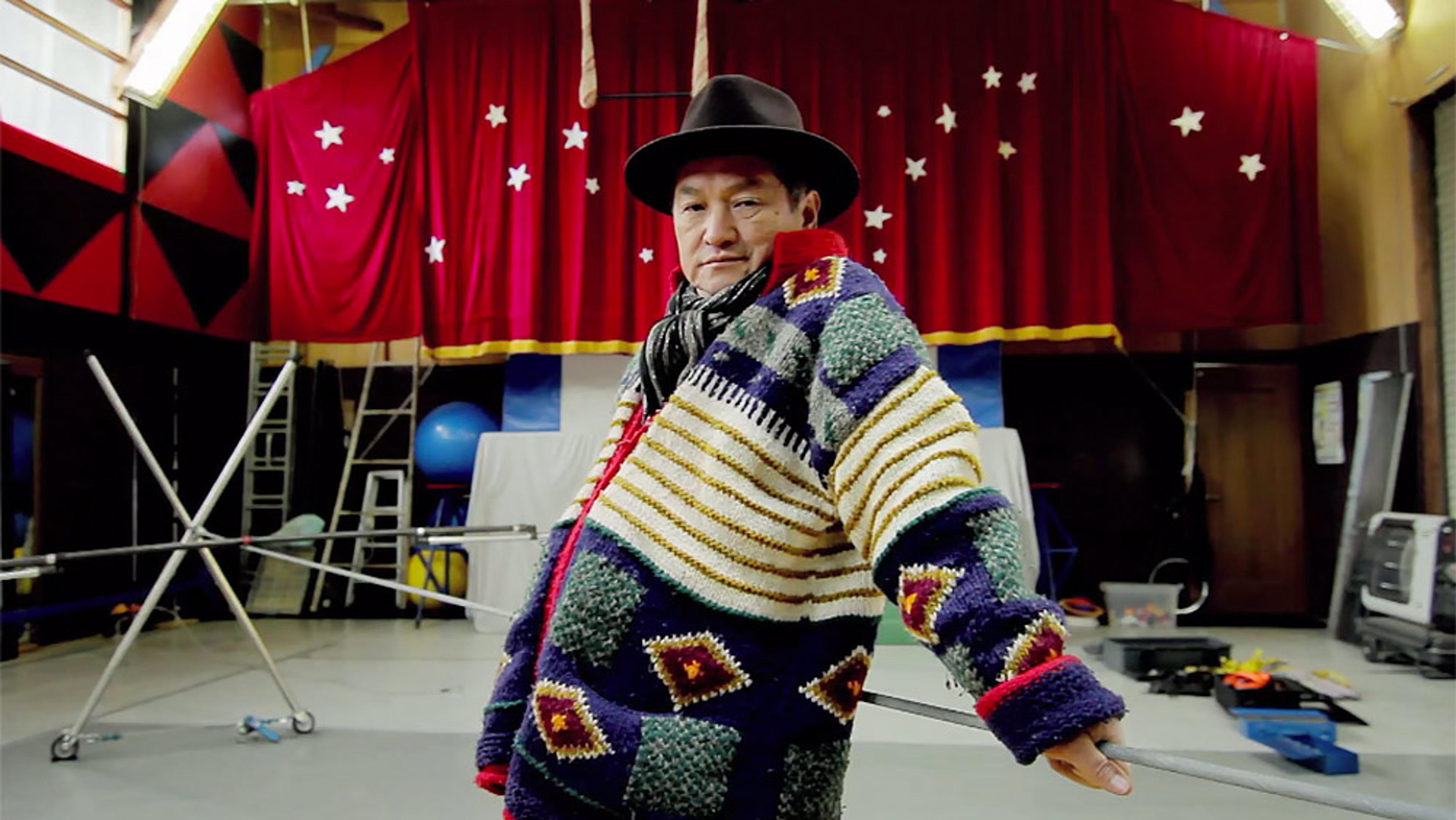 A person in a colourful sweater and hat, standing in a room with red curtains decorated with stars, ladders, and equipment.