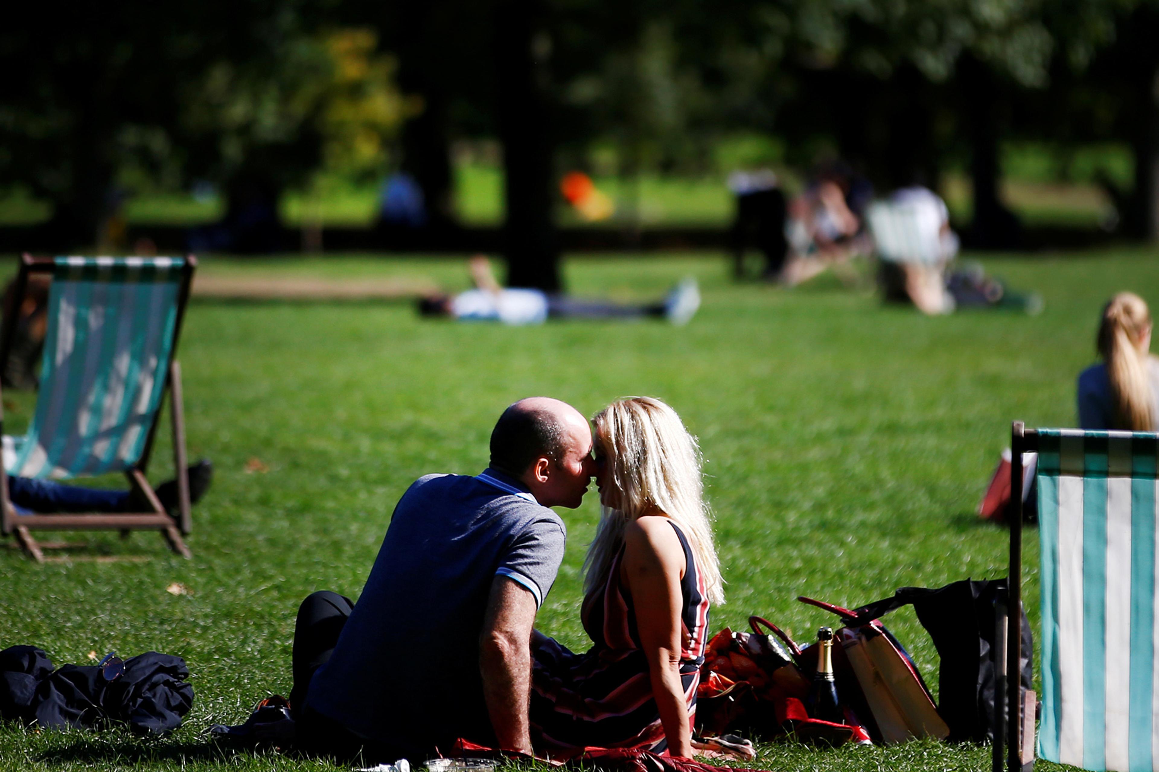 A couple sitting on grass, kissing in a park with deck chairs and people relaxing in the background.