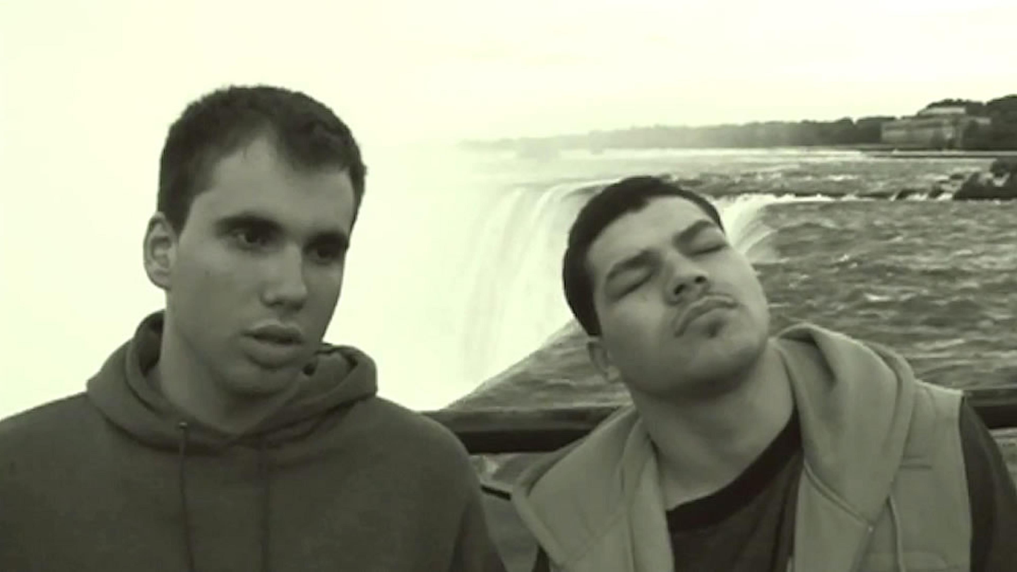 A sepia-toned photo of two men standing in front of a large waterfall, one looking to the side while the other is tilting his head back.