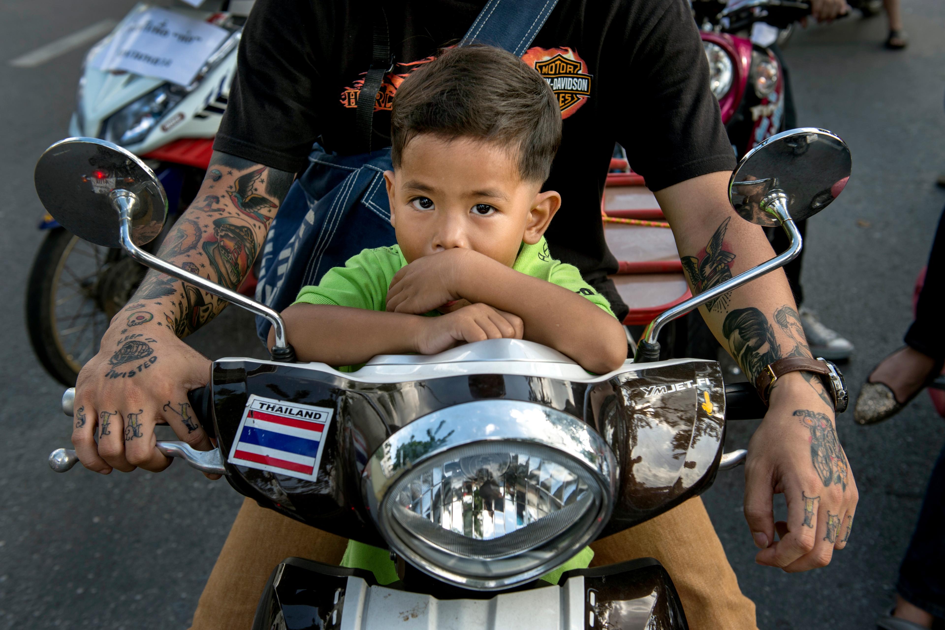 Photo of a child in green on a motorcycle with an adult’s tattooed arms holding the handlebars.