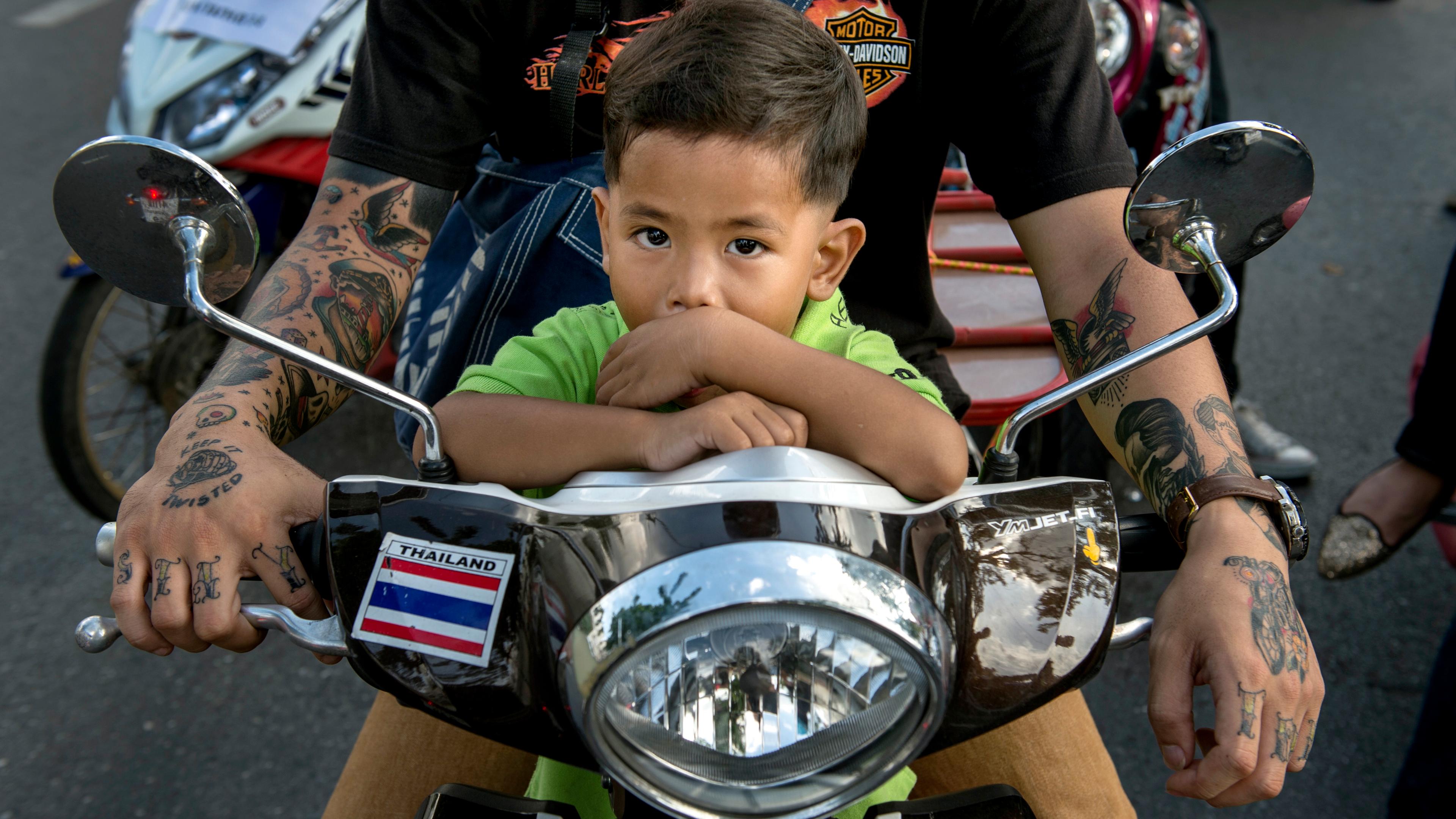 Photo of a child in green on a motorcycle with an adult’s tattooed arms holding the handlebars.