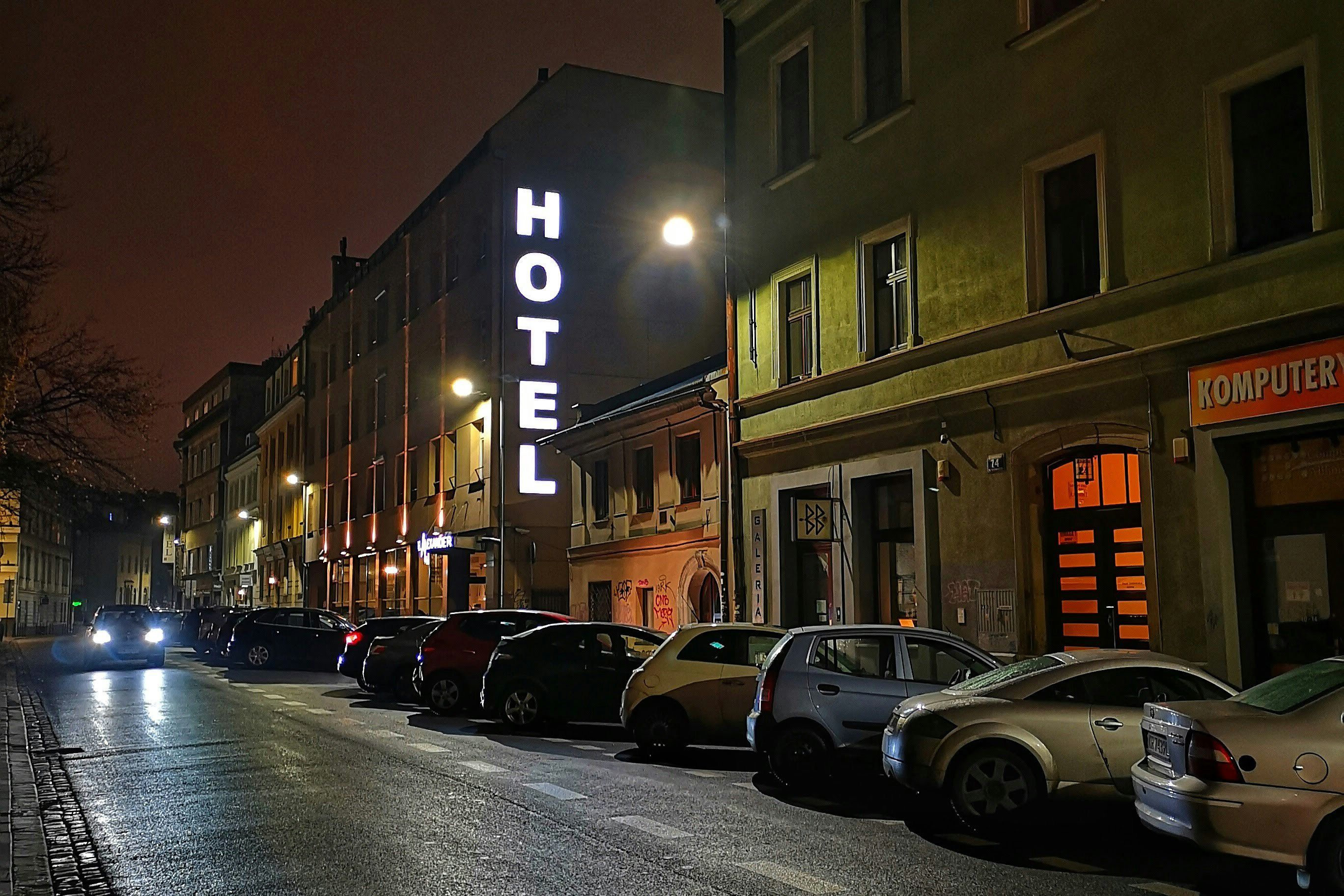 A dimly lit street with parked cars at night. A building has a large, vertical “HOTEL” sign illuminated.