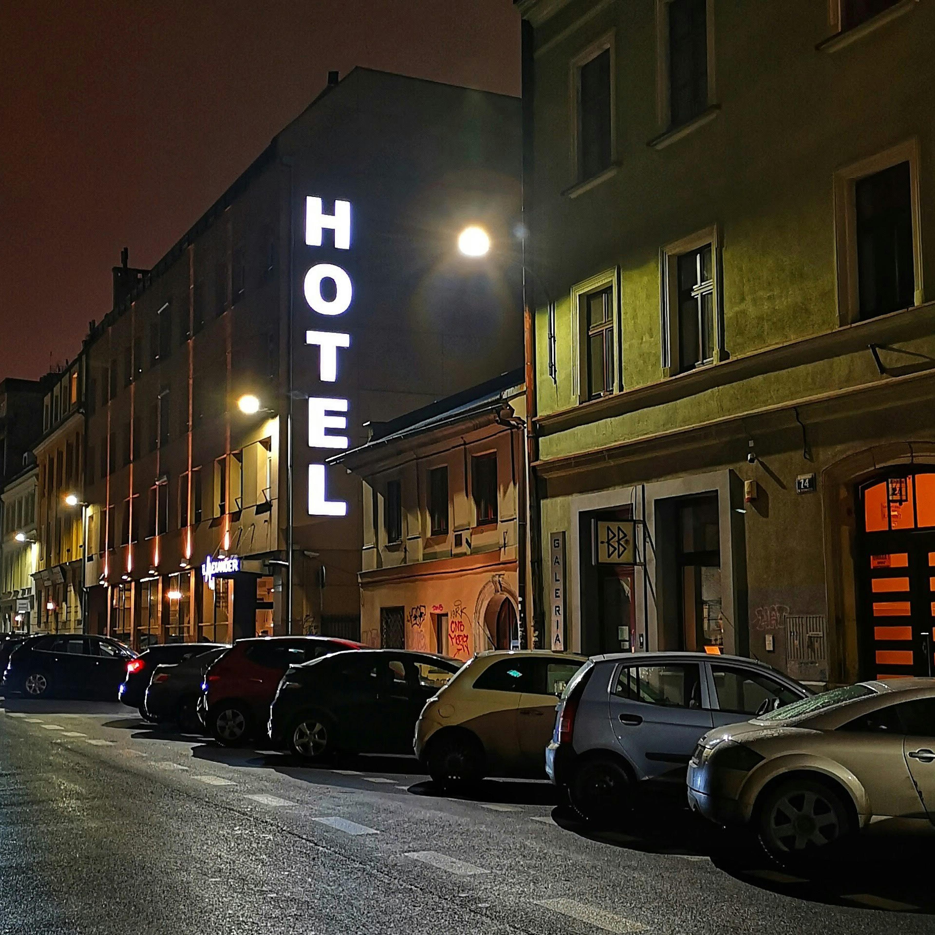 A dimly lit street with parked cars at night. A building has a large, vertical “HOTEL” sign illuminated.