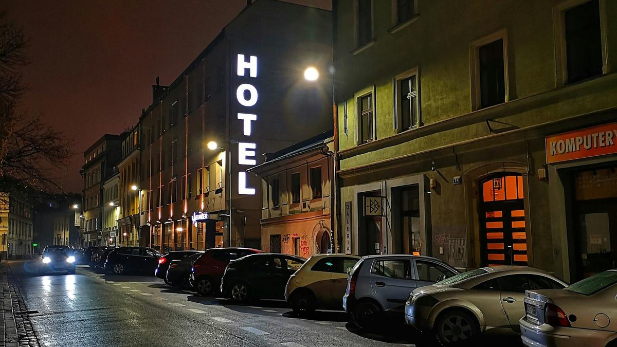 A dimly lit street with parked cars at night. A building has a large, vertical “HOTEL” sign illuminated.