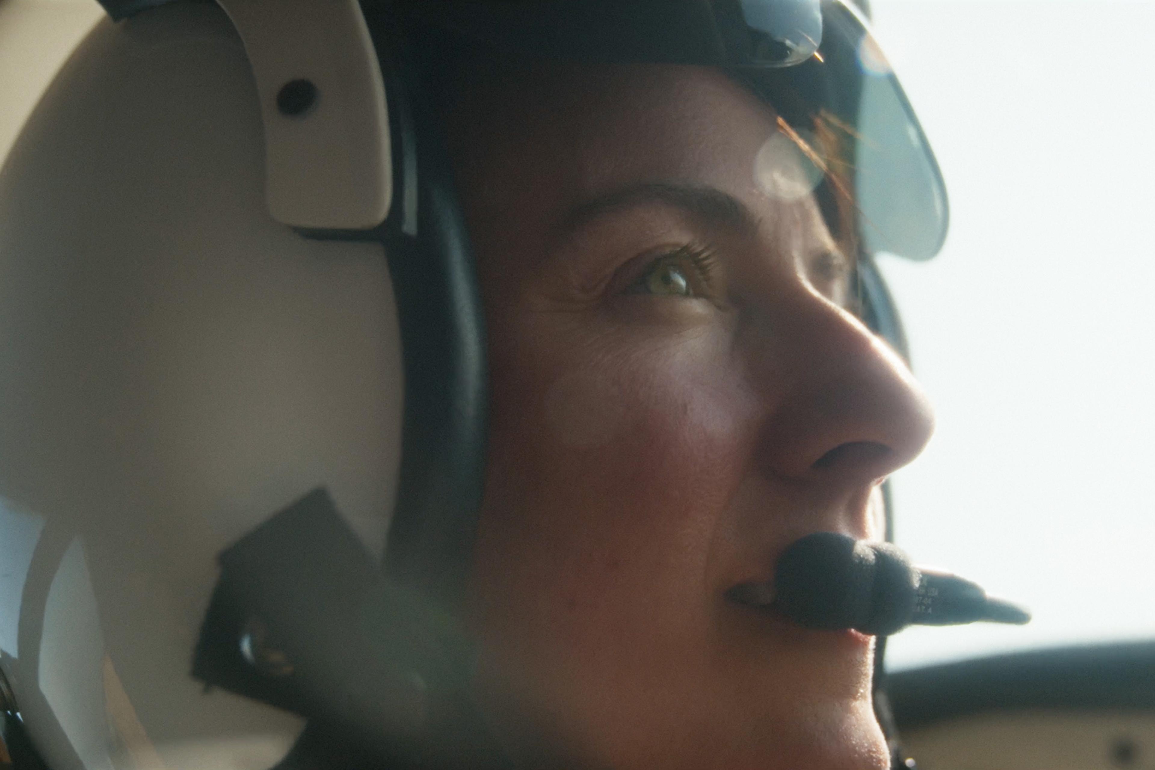 Close-up of a woman wearing a flight helmet with a microphone, looking ahead with a focused expression.