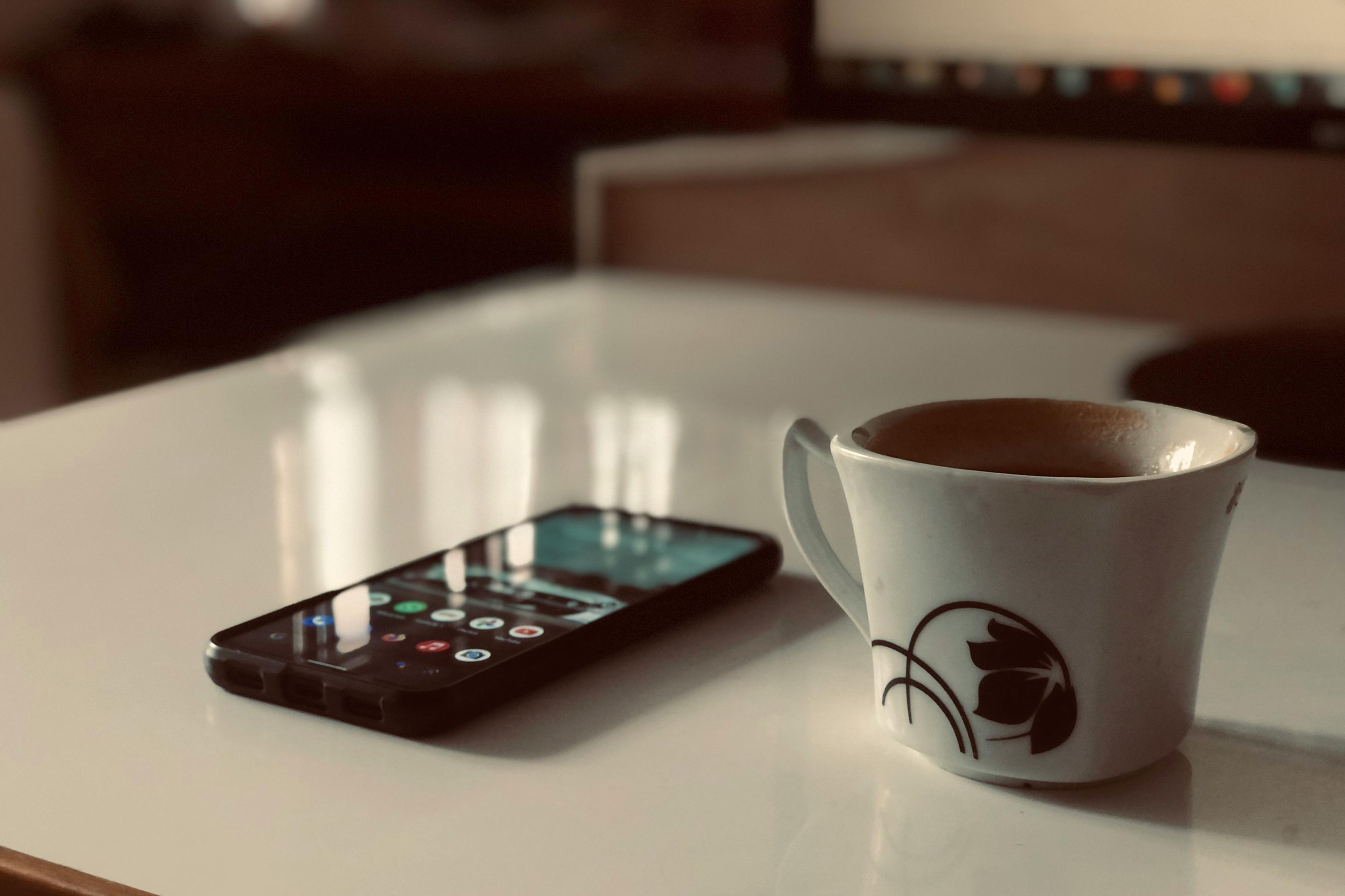 A white table with a smartphone and a white mug of coffee with a floral design, in a softly lit room.