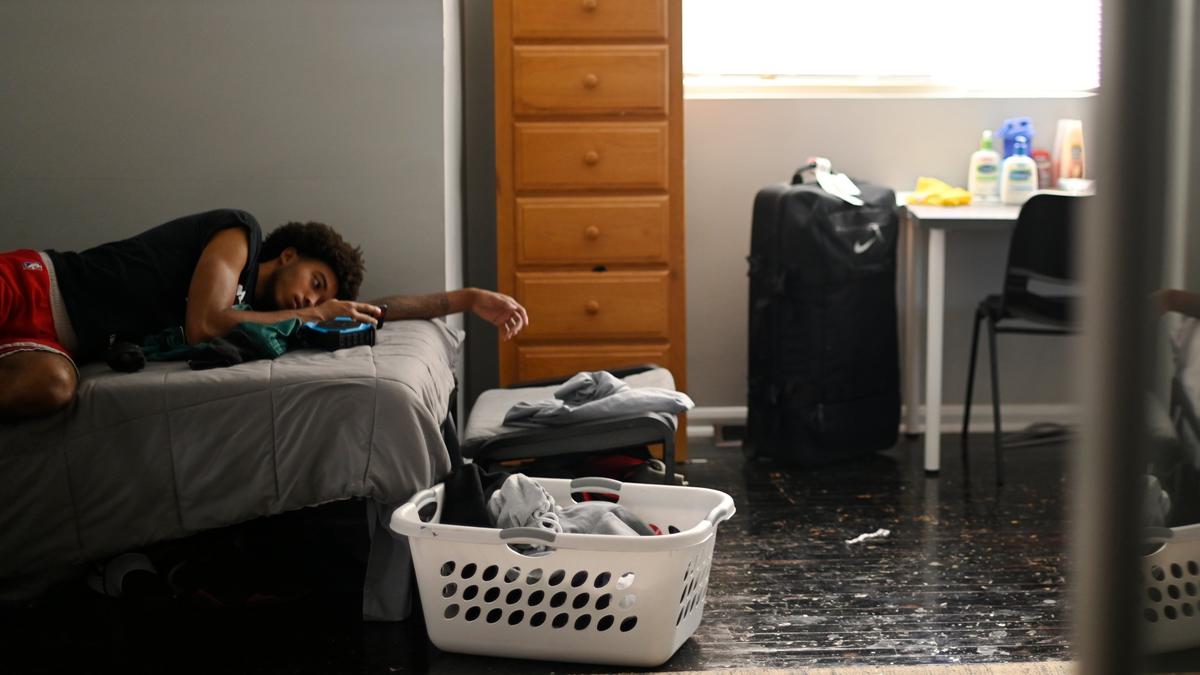 A young person lying on a bed in a cluttered room with a suitcase, laundry basket, chest of drawers and desk, illuminated by window.