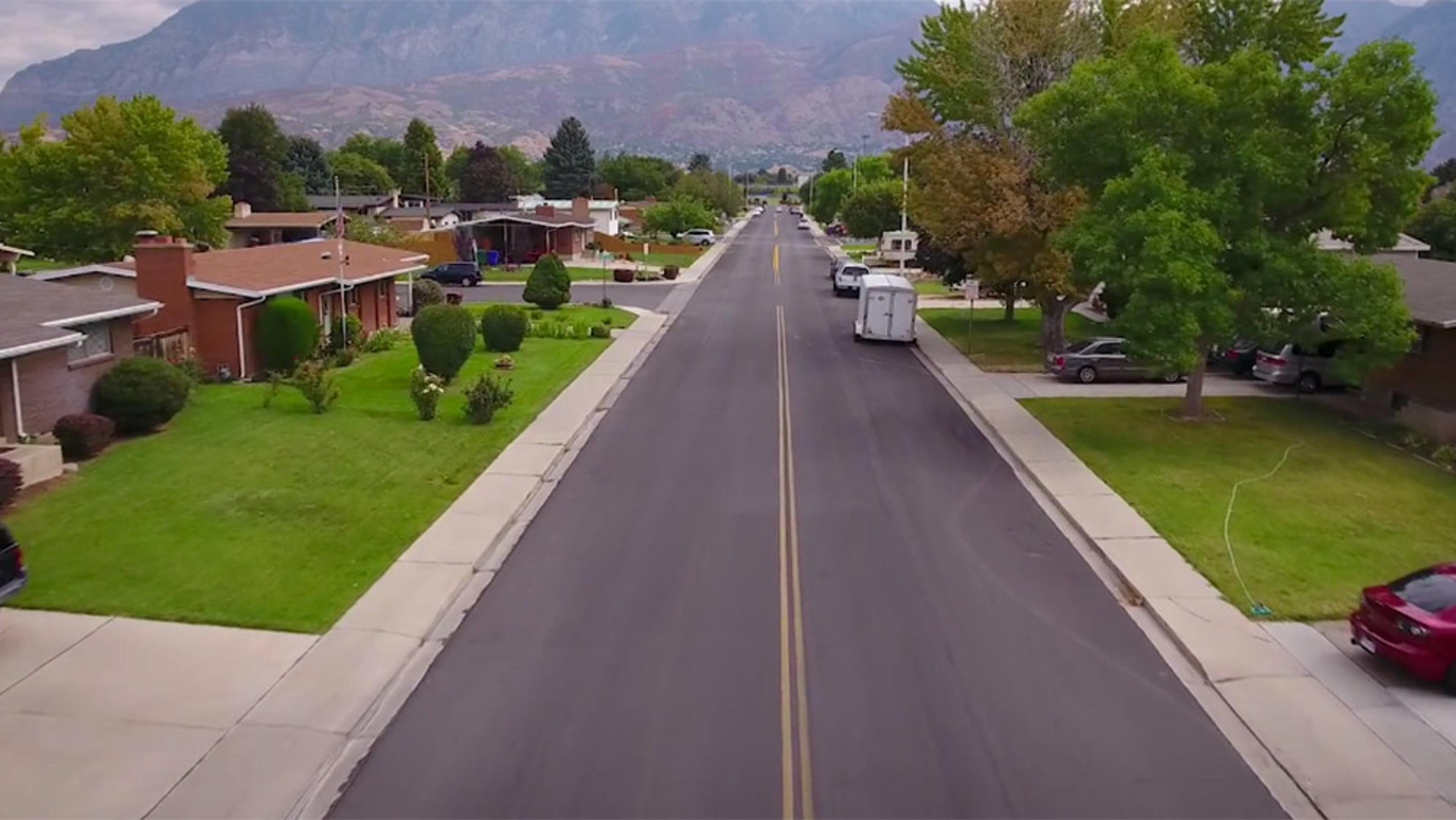 A suburban street with houses, trees, grass, and cars, and a mountain range in the background, taken from an aerial view.