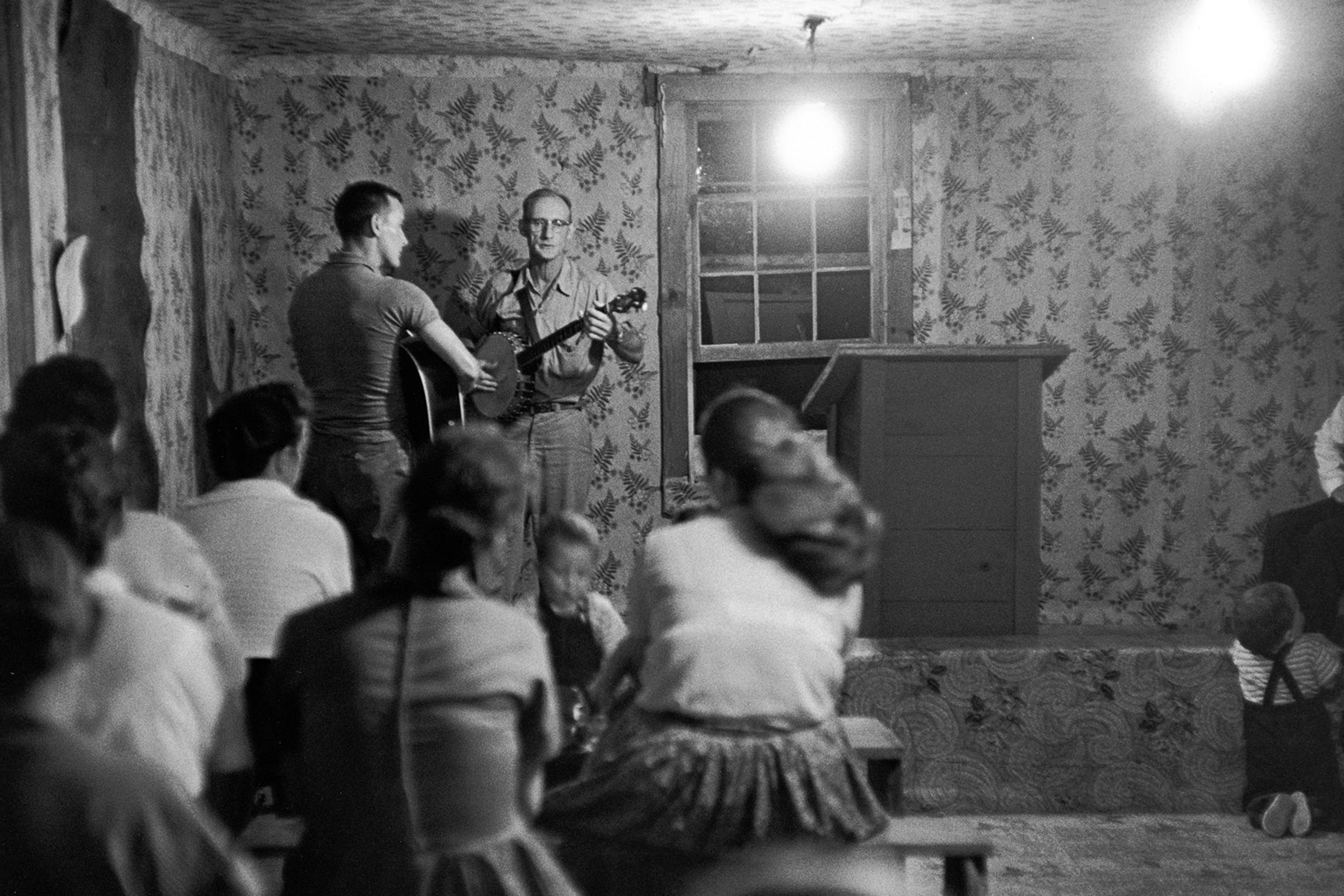 Black-and-white photo of people watching two musicians on stage in a small, wallpapered room with a single light bulb hanging.