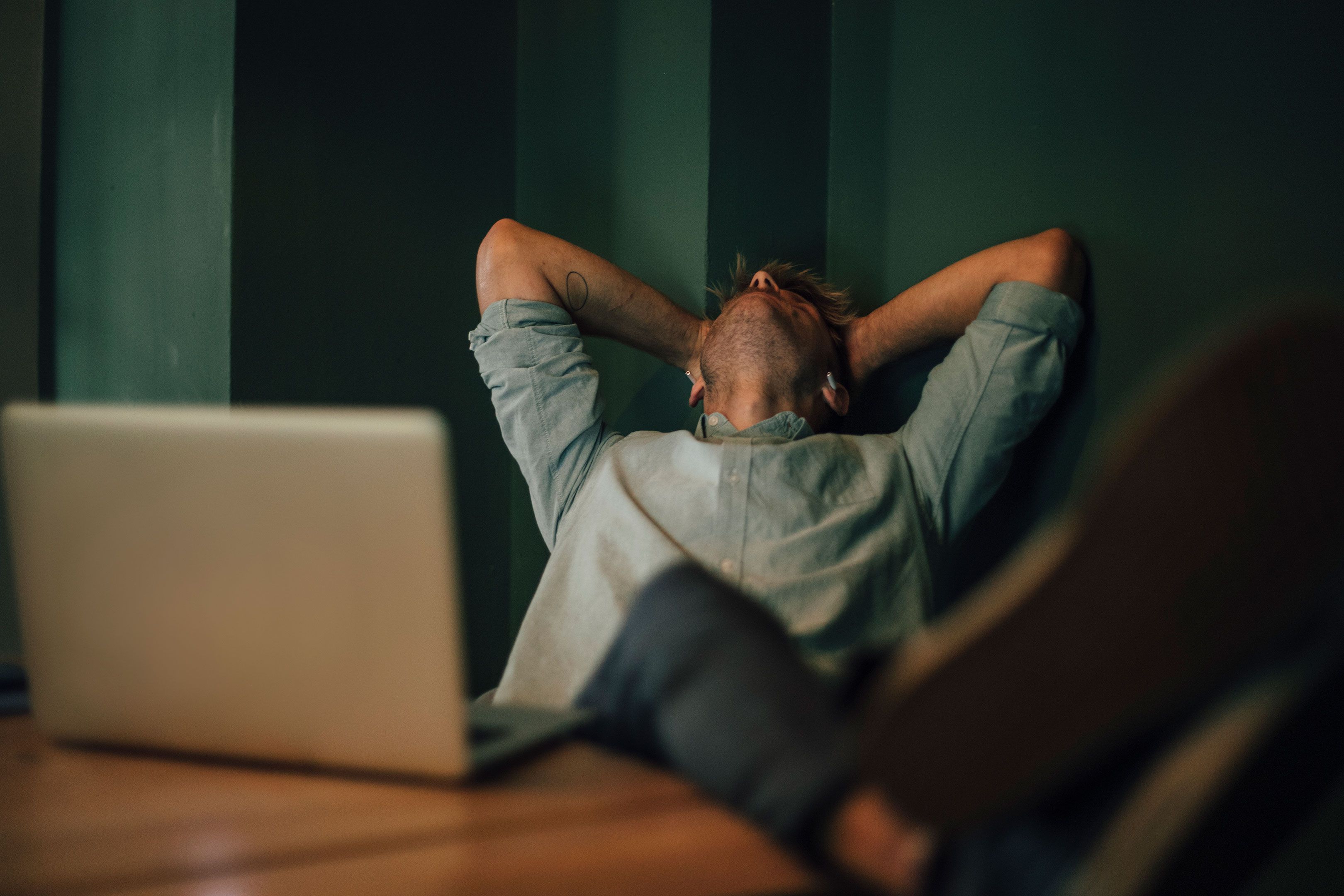 Photo of a person leaning back with hands on head at a desk, laptop open, with feet up on the desk, green background.