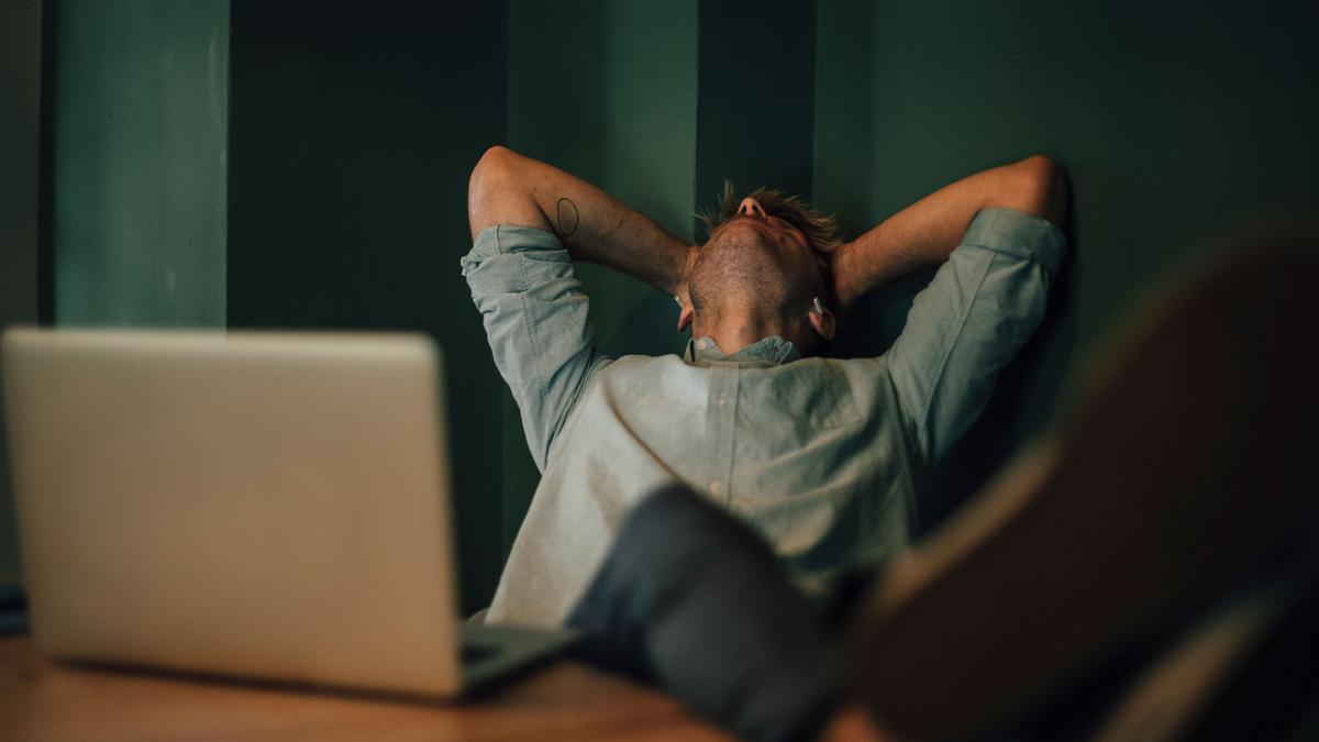 Photo of a person leaning back with hands on head at a desk, laptop open, with feet up on the desk, green background.